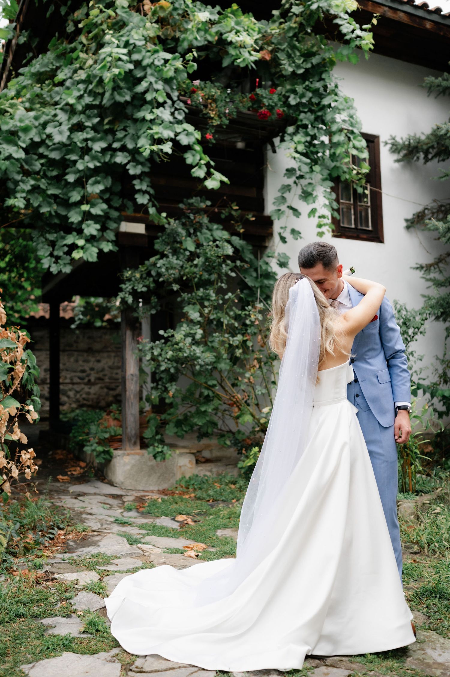 A couple in wedding attire share romantic moments in a lush garden setting with stone paths and greenery.