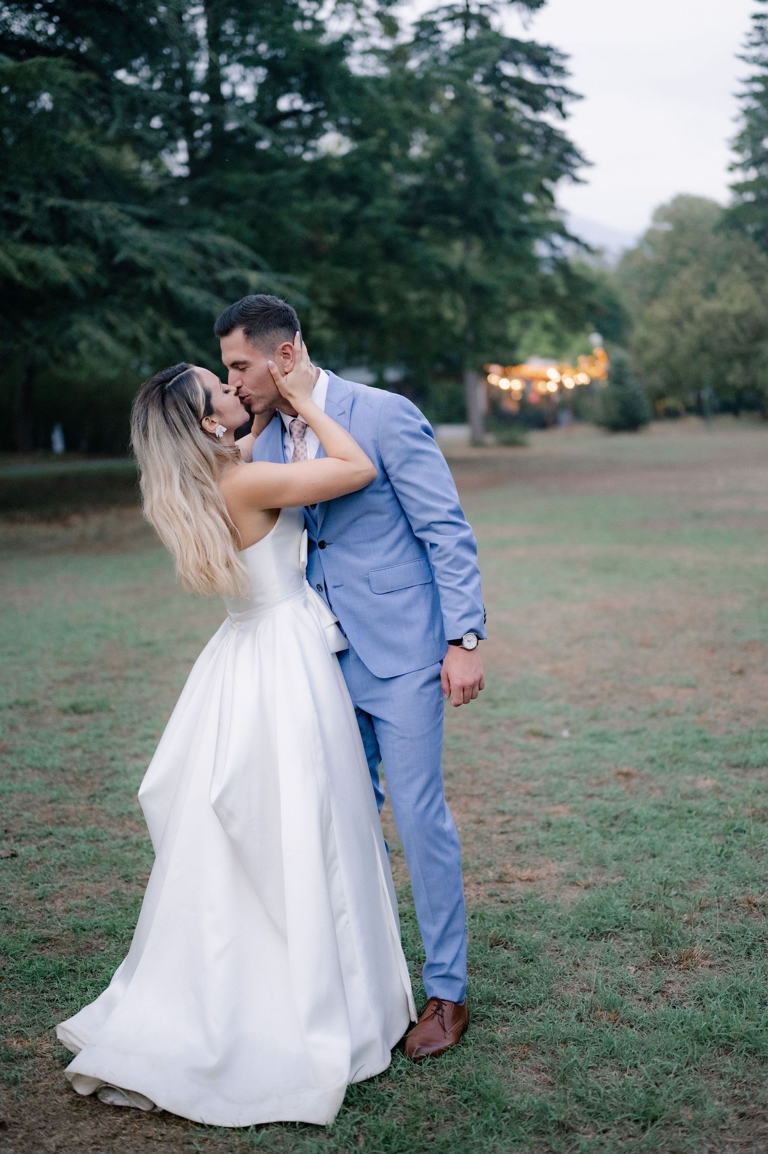 A romantic couple shares intimate moments in a grassy field surrounded by evergreen trees at sunset.