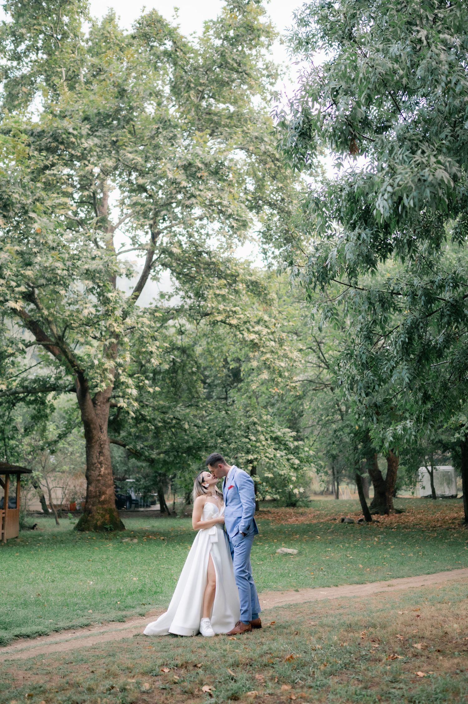 A couple shares a romantic moment under tall trees in a serene park setting during their wedding photoshoot.