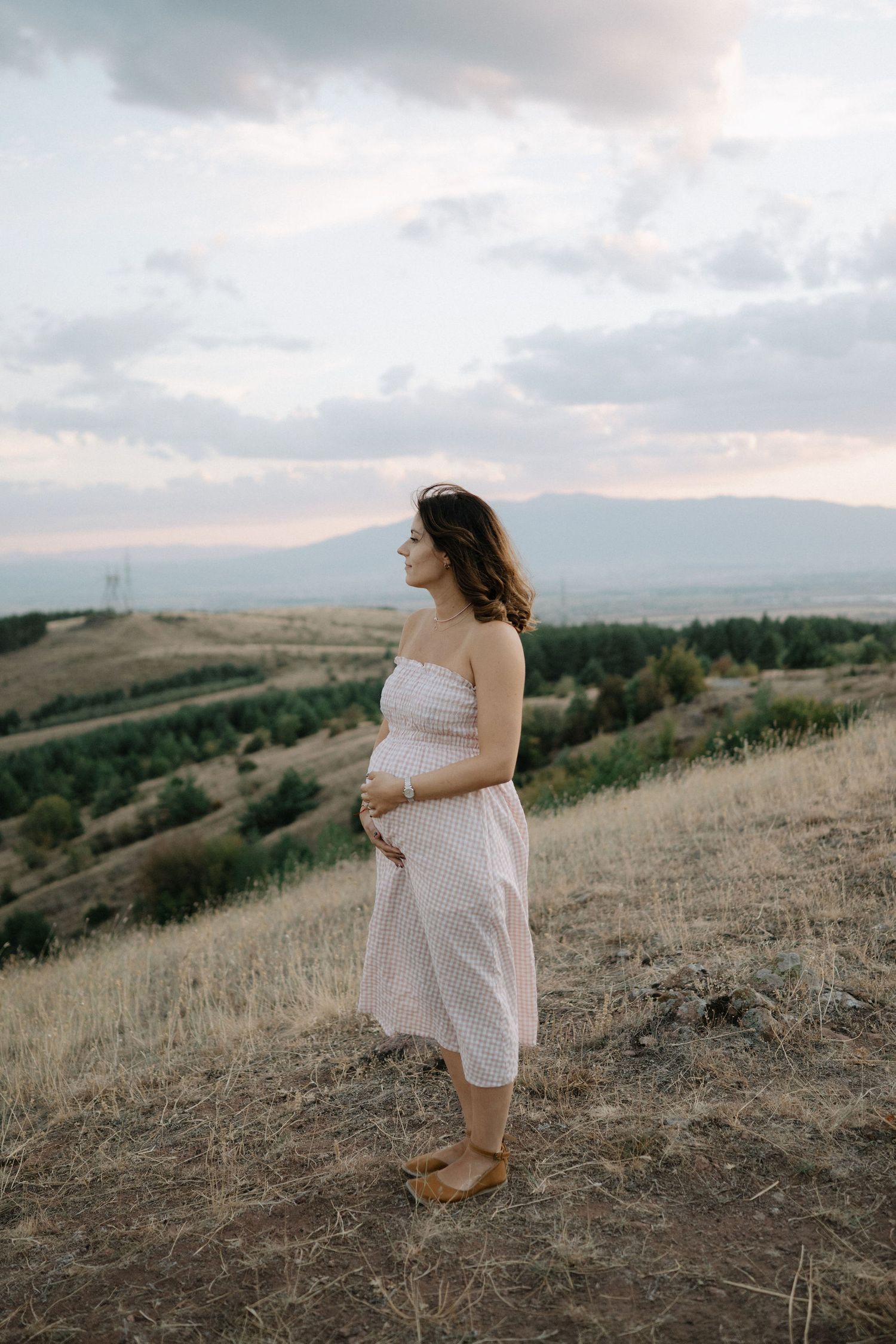 A pregnant woman in a white dress stands on a hilltop against a cloudy sky at sunset in a scenic landscape.