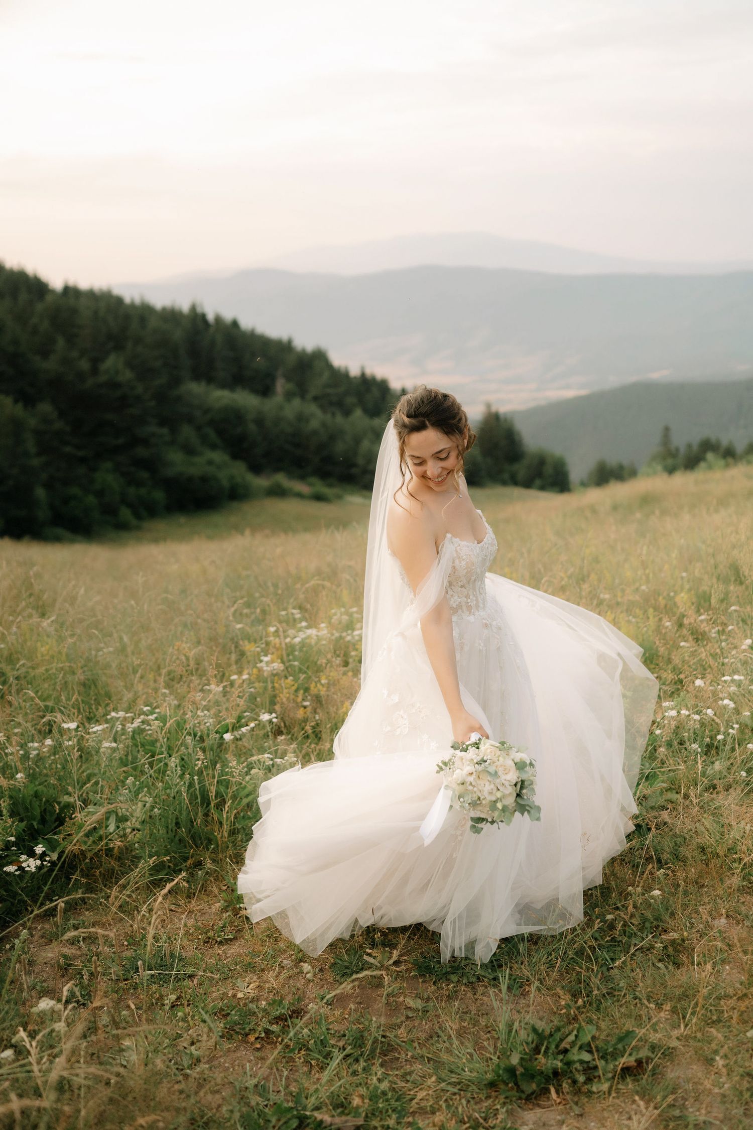 A bride in a flowing white dress twirls gracefully in a mountain meadow filled with wildflowers at sunset.