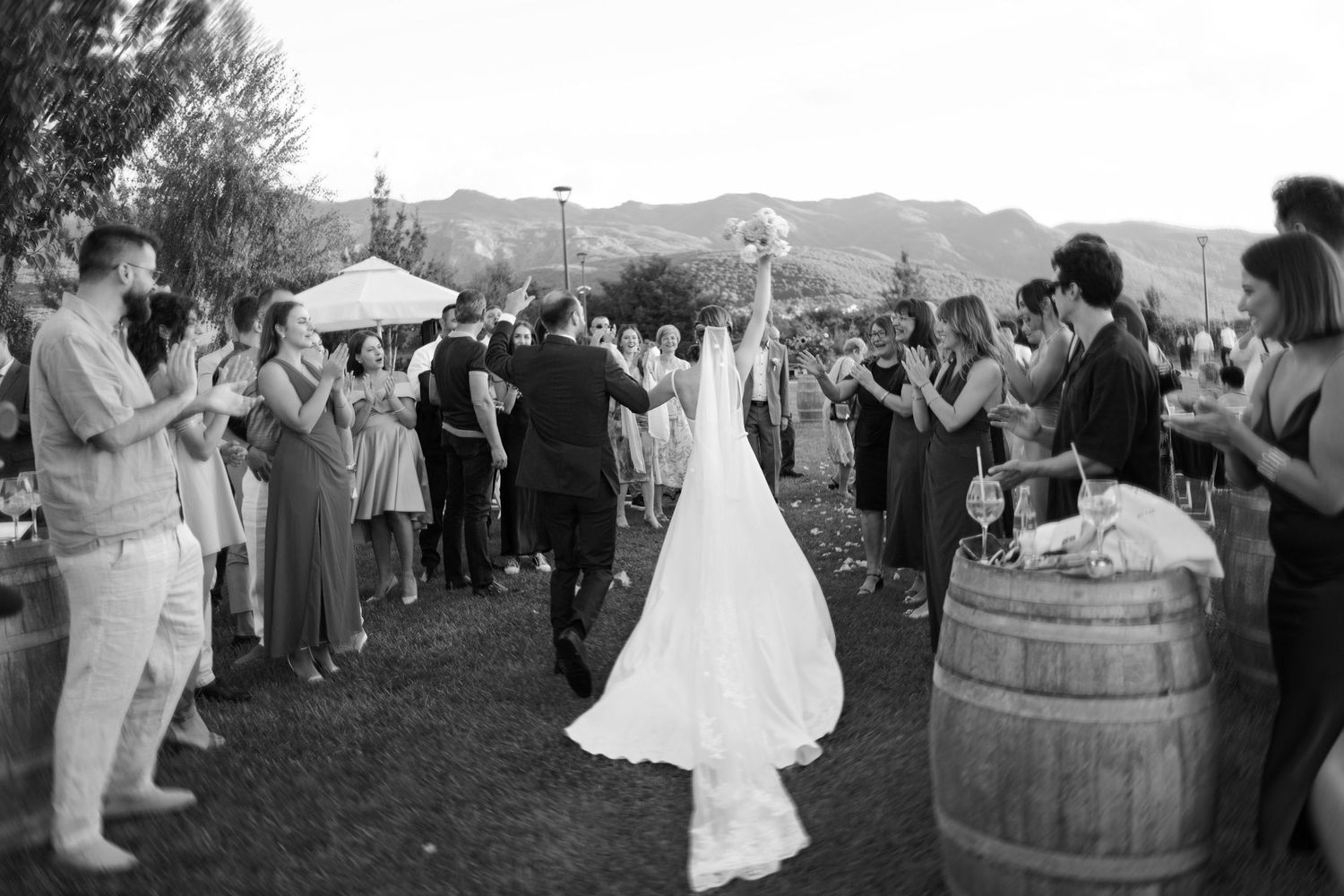 Black and white photo of an outdoor wedding celebration with guests forming a pathway on a lawn with mountains in background.