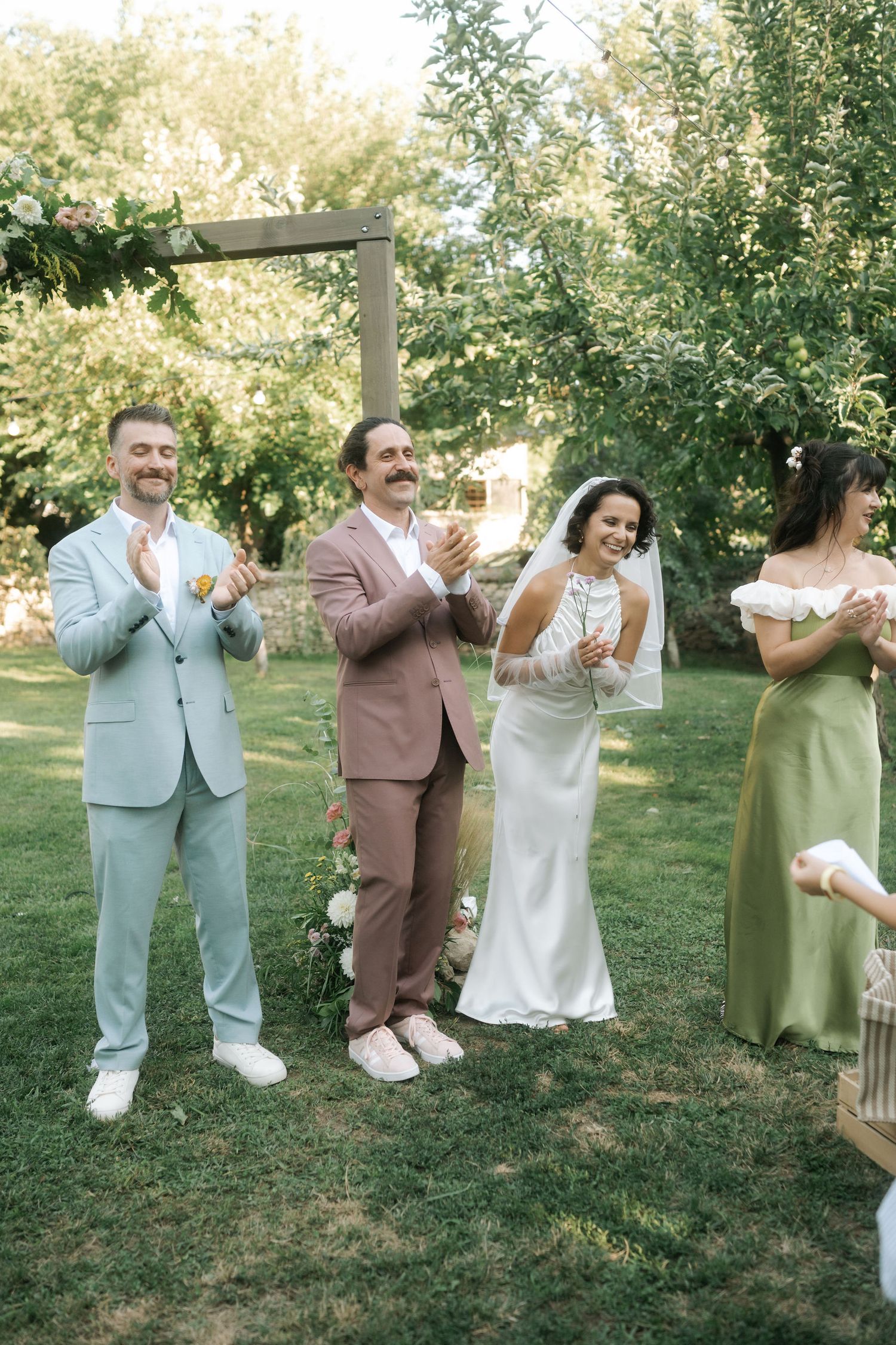 Wedding party members perform a fun dance sequence in an outdoor garden setting under green trees.
