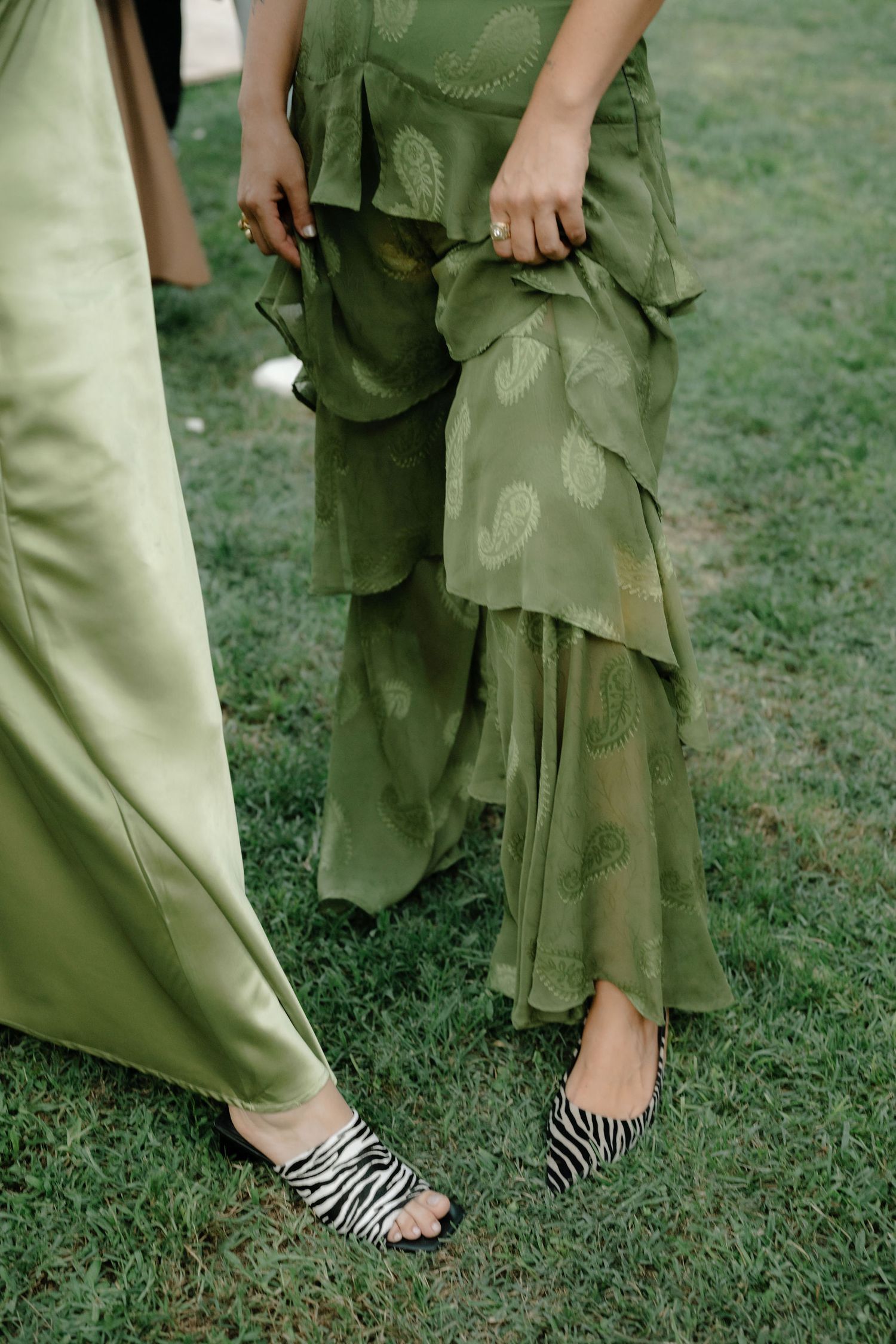 People wearing green pants and zebra print sandals stand together on grass.