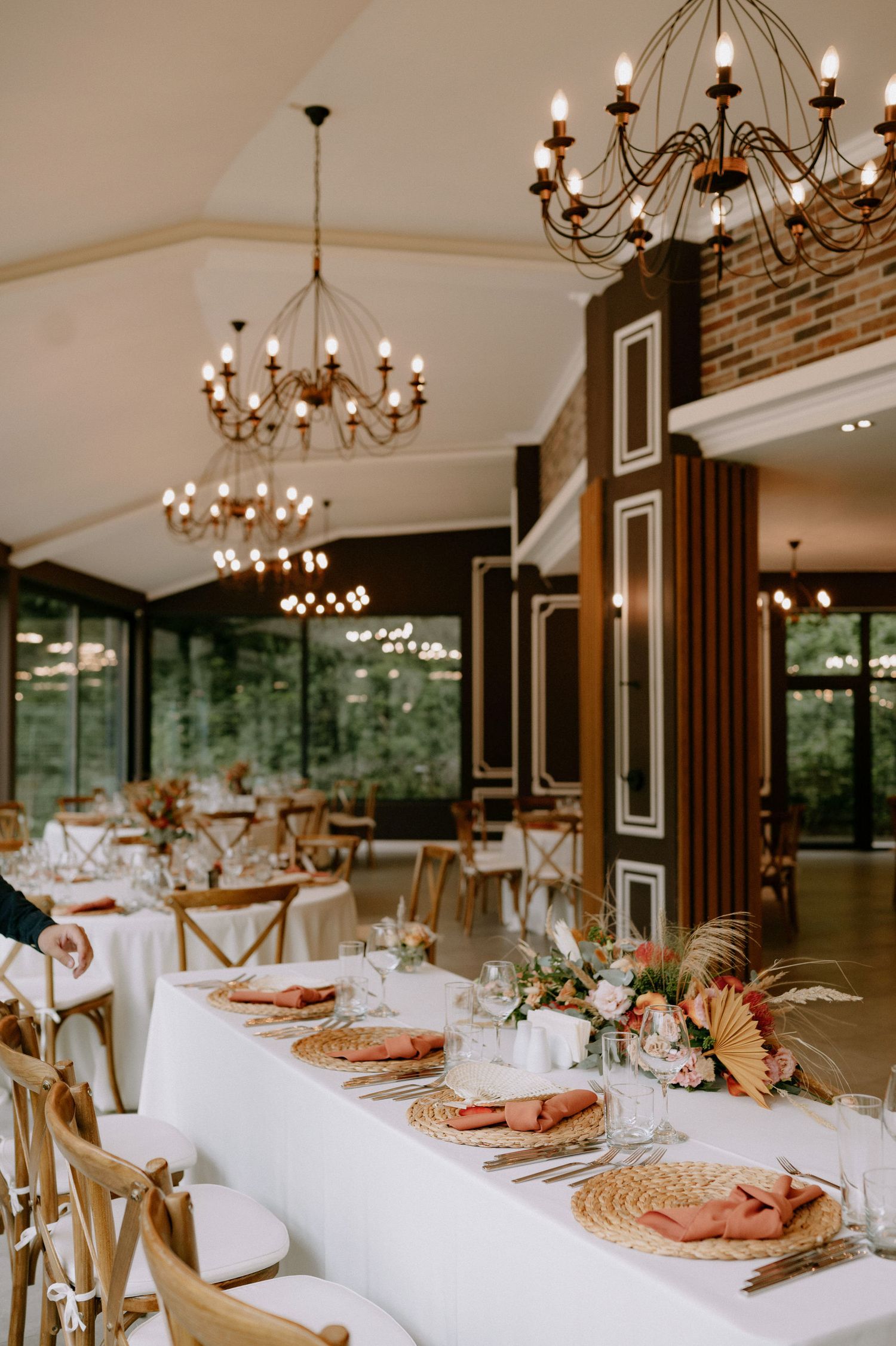Elegant wedding reception venue with long tables set with woven placemats and chandeliers in a rustic-modern space.