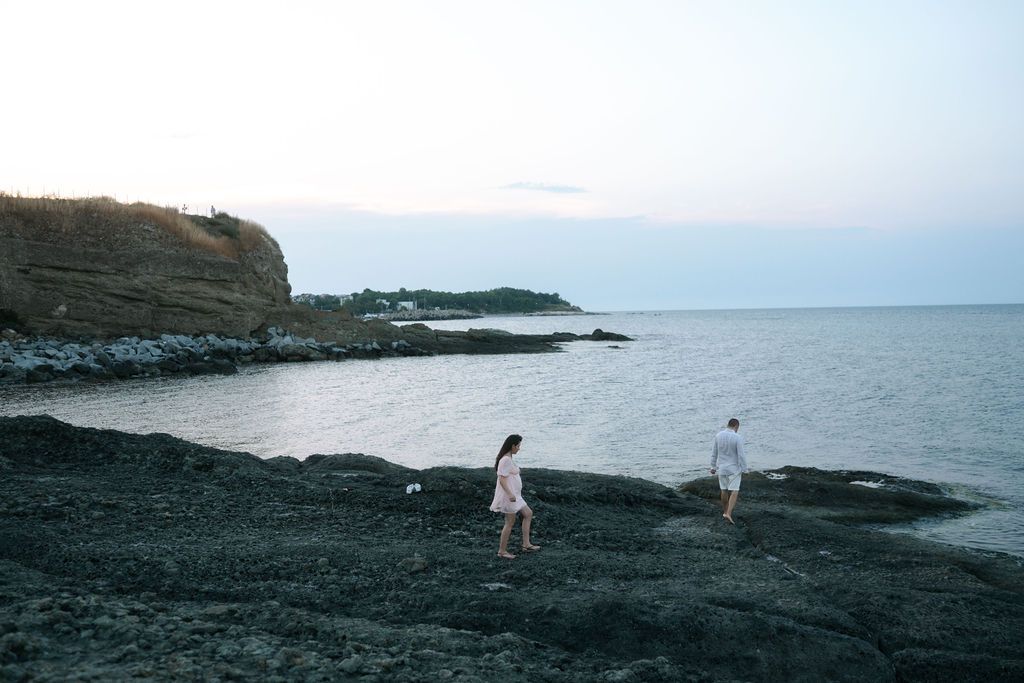 Two people explore a rocky volcanic beach with cliffs and ocean views at dusk.