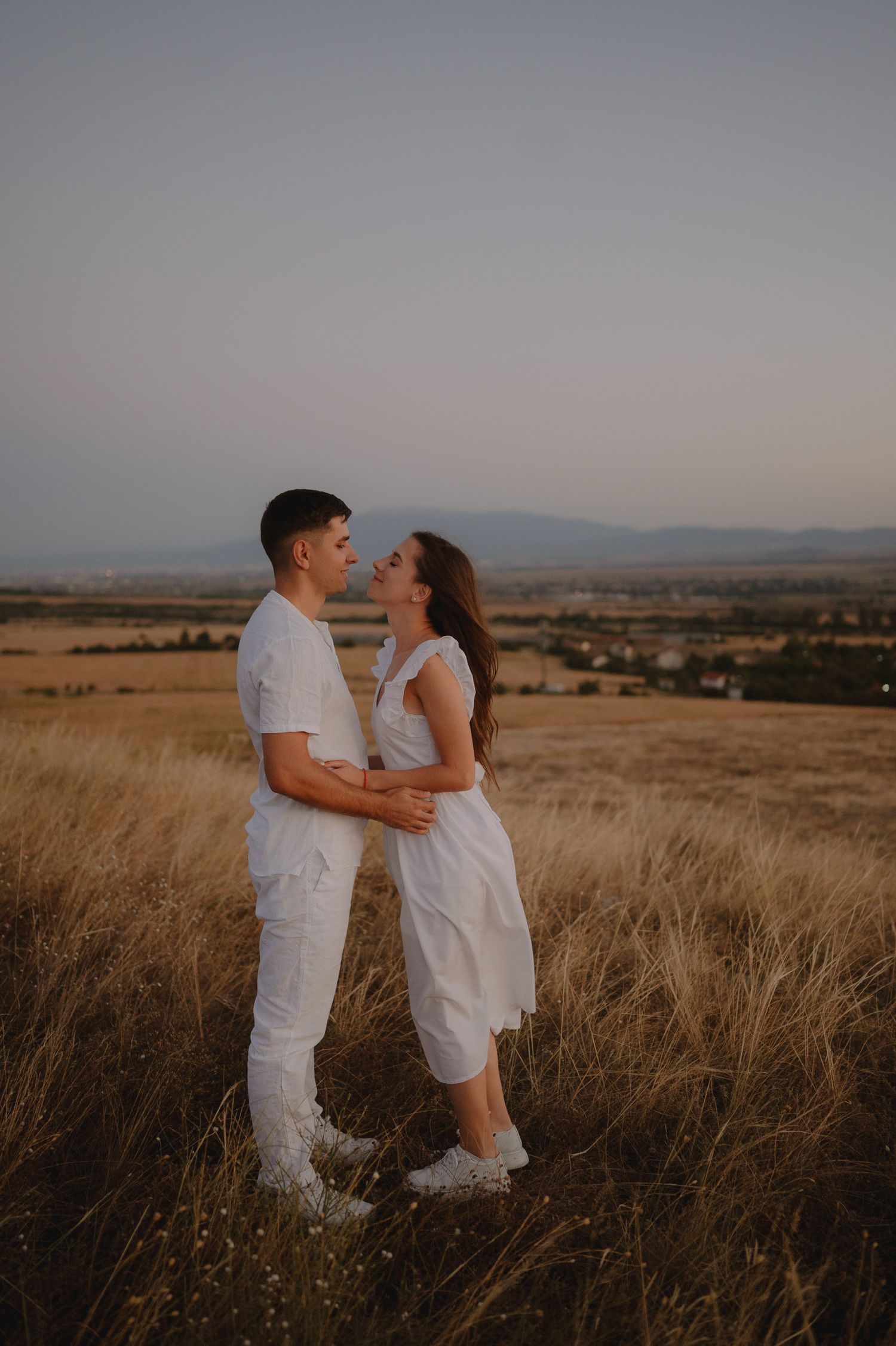 A couple in white outfits embraces lovingly in a golden field at sunset with mountains in the distance.