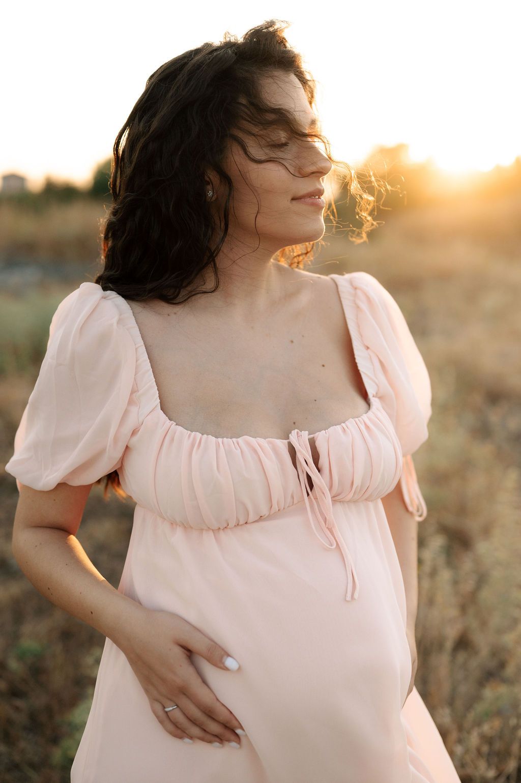 Woman in romantic white dress poses in a field during golden hour with sunlit silhouette.