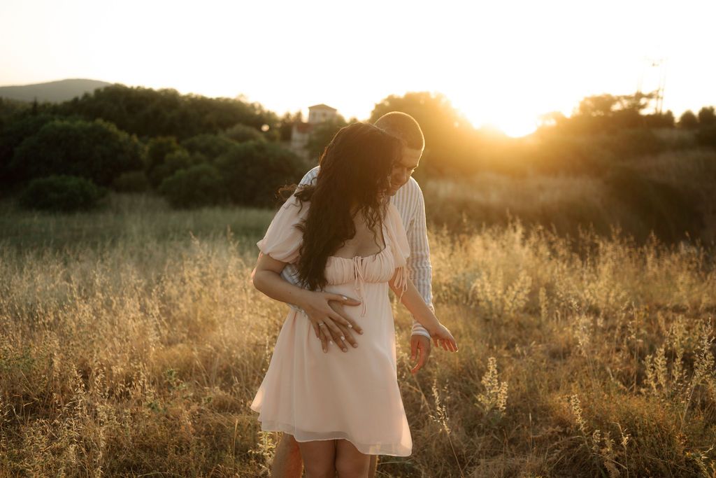 Golden sunlight illuminates a romantic couple in a white dress and suit walking through a grassy field at sunset.