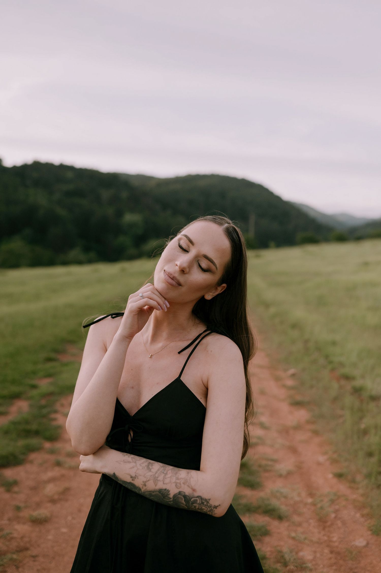 A person in a black top poses expressively against a scenic mountain landscape on a dirt path.