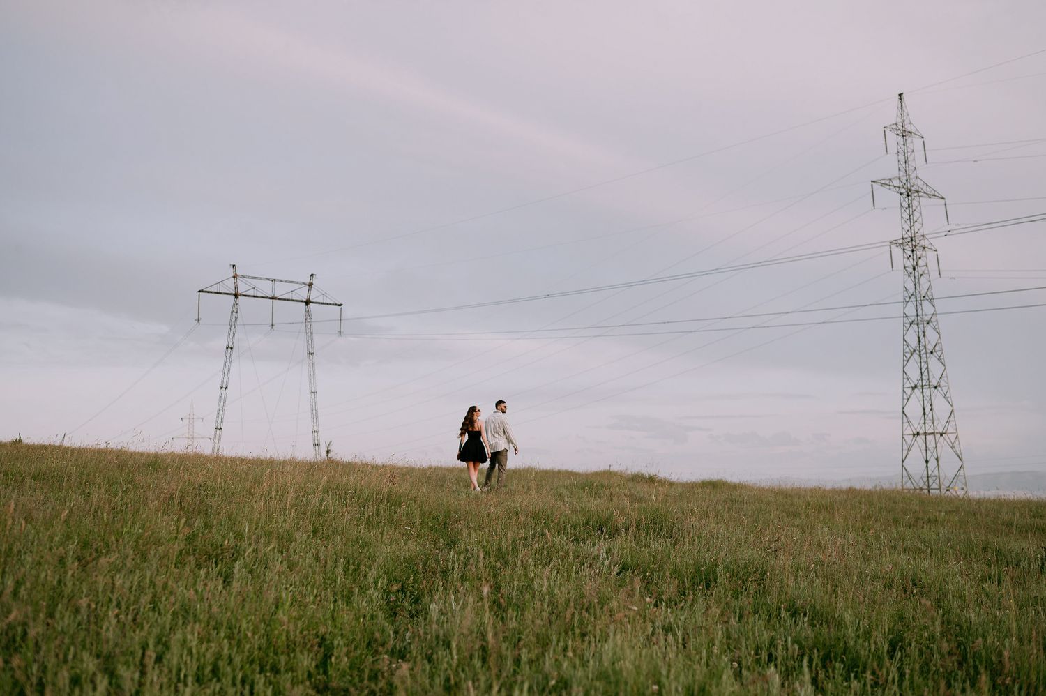 Silhouetted figures walk along a grassy hilltop beneath towering electrical transmission towers and power lines.
