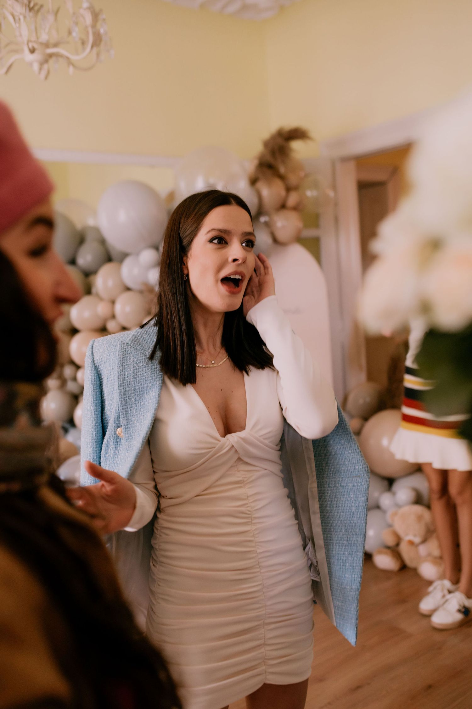 A woman in a white dress and light blue cardigan poses expressively in a room with plush decor.
