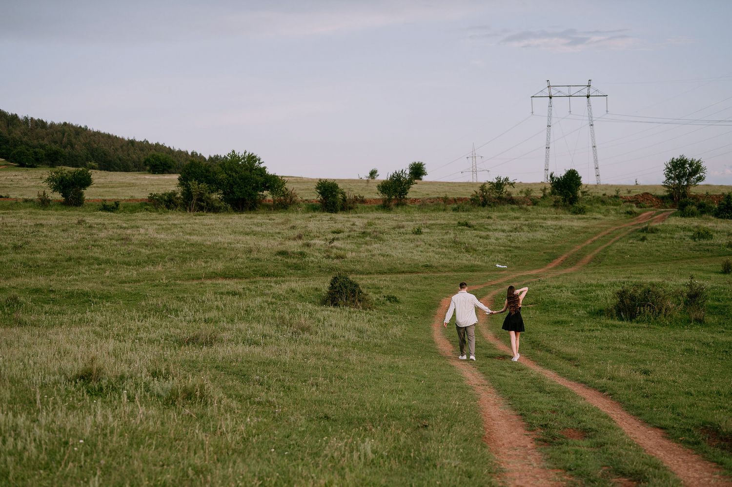 Two people walk along a dirt path through rolling green prairie grasslands under power lines.