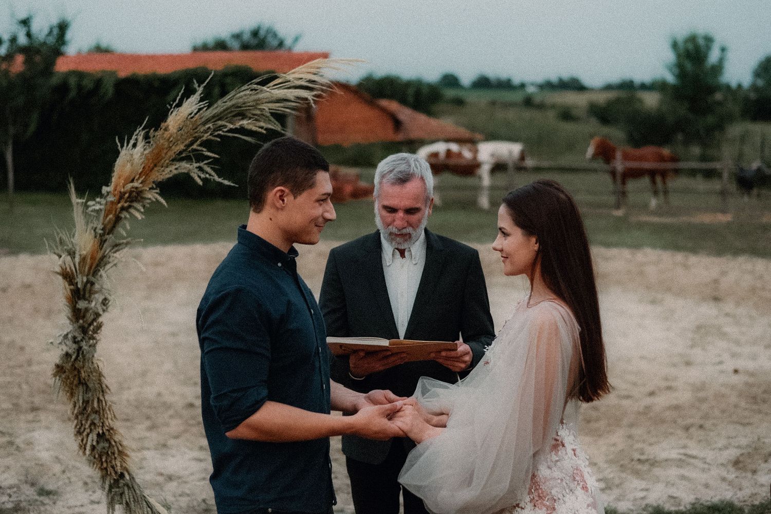 A couple stands together during an outdoor wedding ceremony on a sandy beach at sunset.