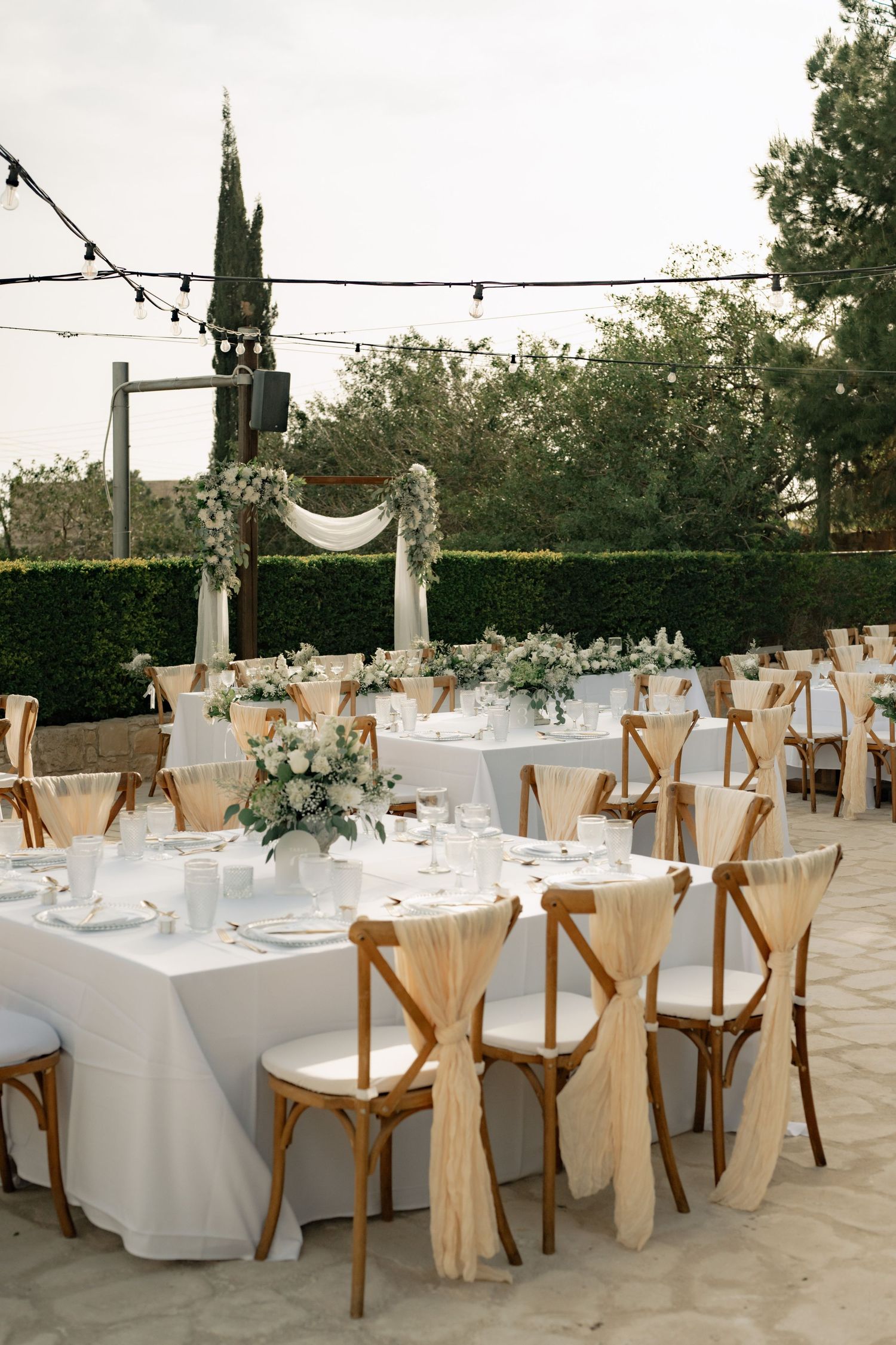 Elegant outdoor wedding reception with wooden cross-back chairs, white tablecloths, floral centerpieces, and string lights.
