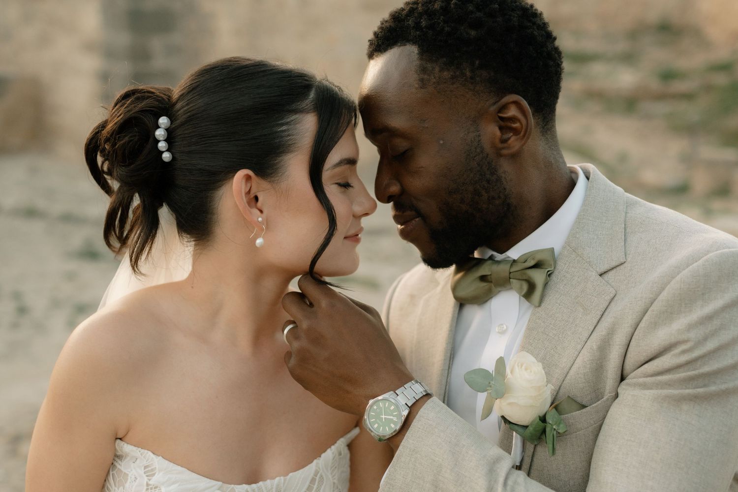 Interracial couple in wedding attire share an intimate moment, groom in beige suit with boutonniere, bride in white dress.