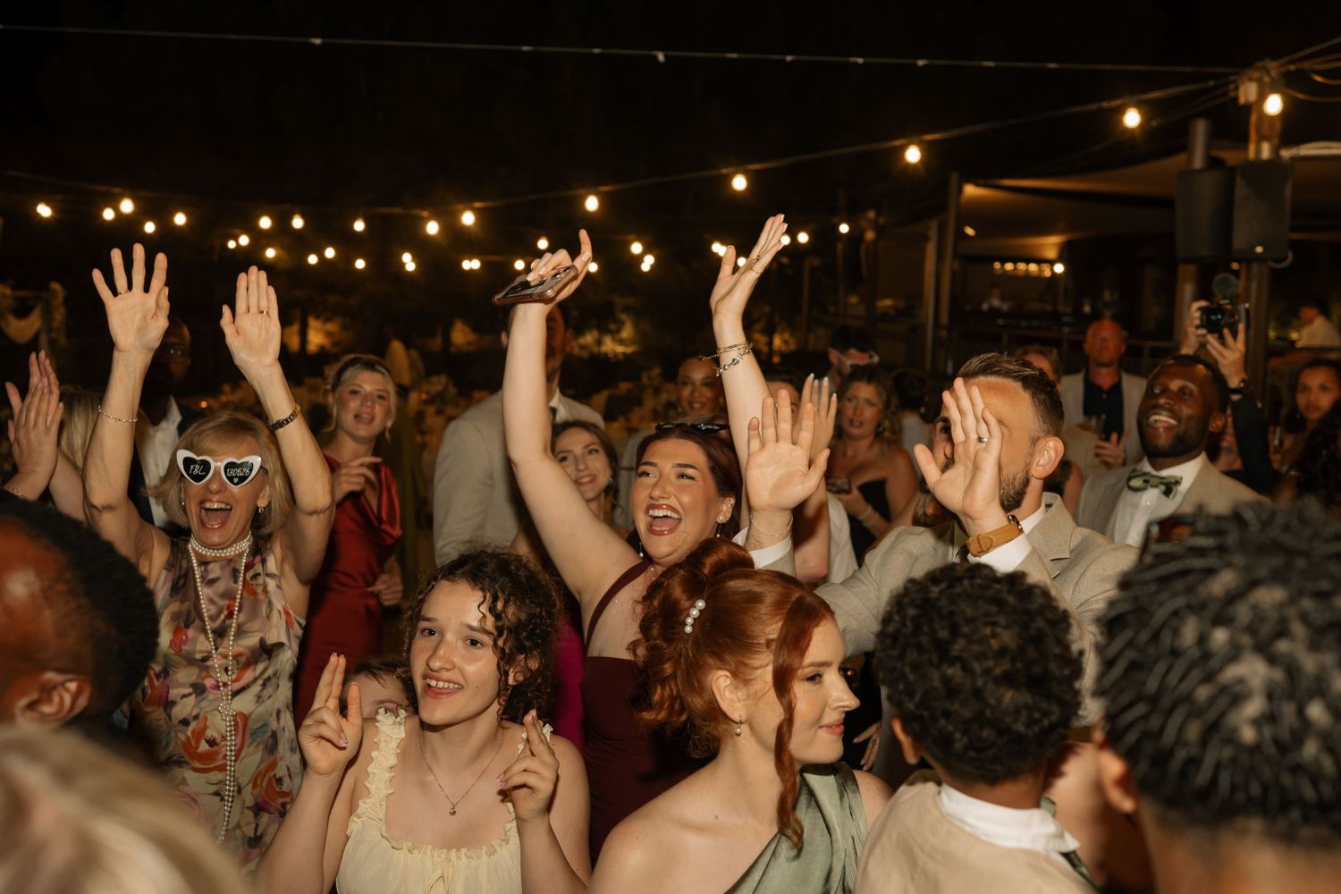Joyful wedding reception guests dancing and celebrating under warm string lights at an outdoor evening party.
