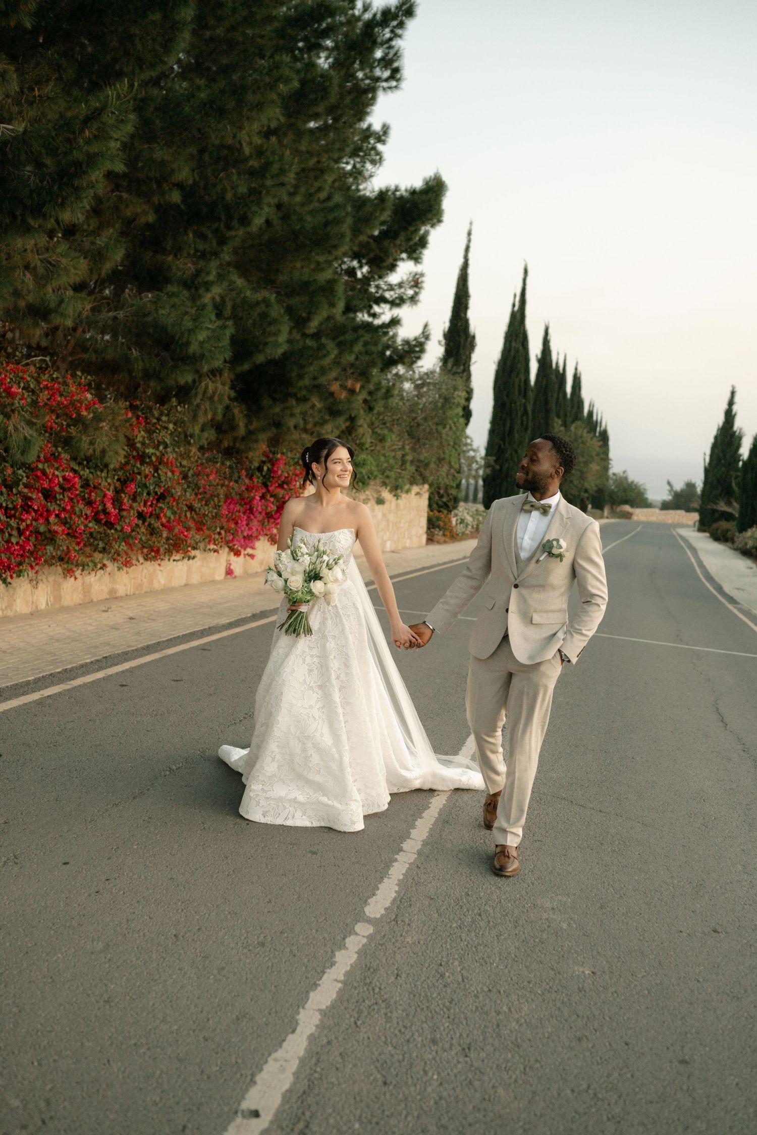 Bride and groom walk hand-in-hand down a scenic road lined with cypress trees and red flowers on their wedding day.