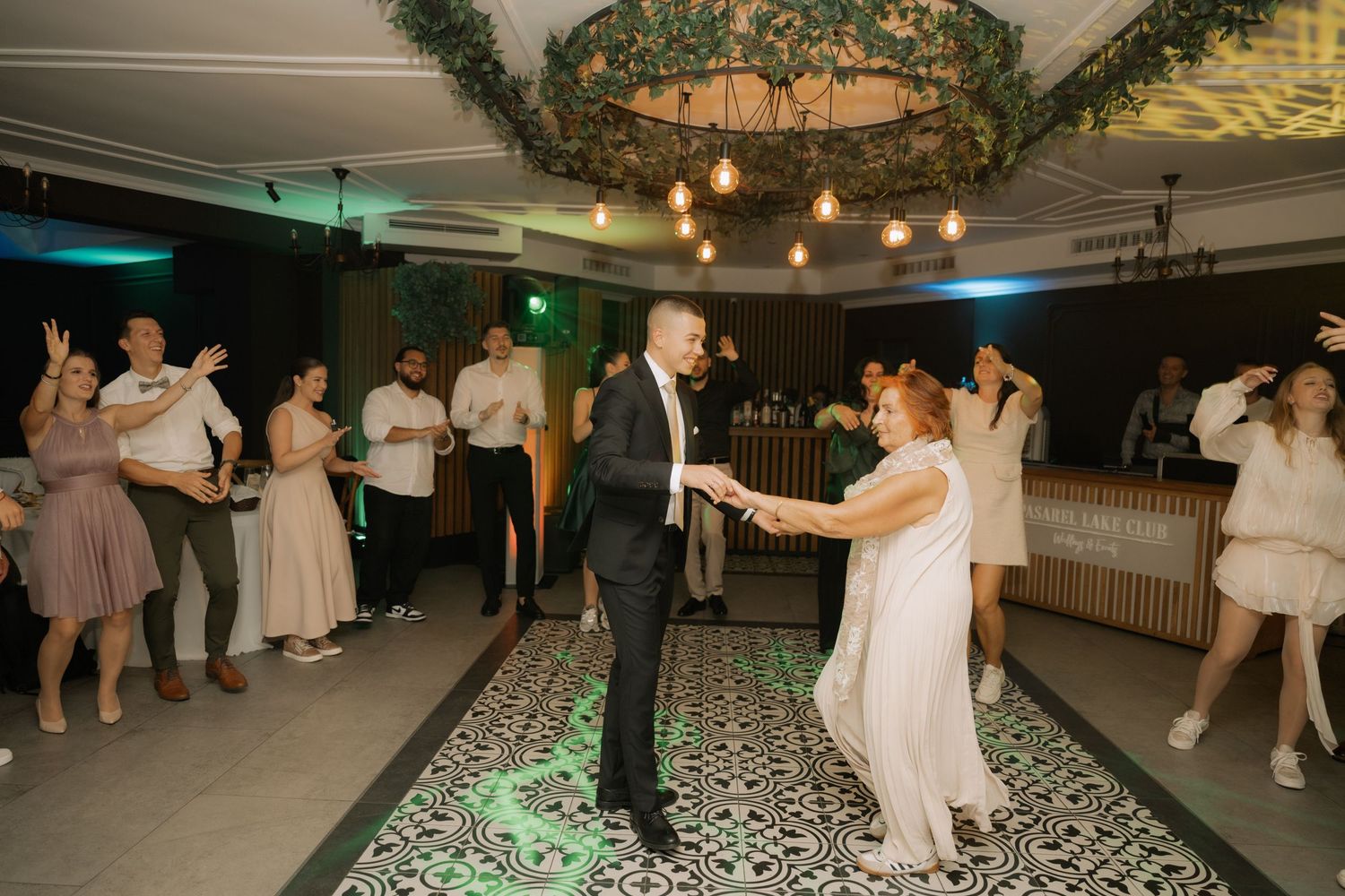 Wedding guests dance and celebrate on a black and white patterned dance floor under warm string lights at an evening reception.