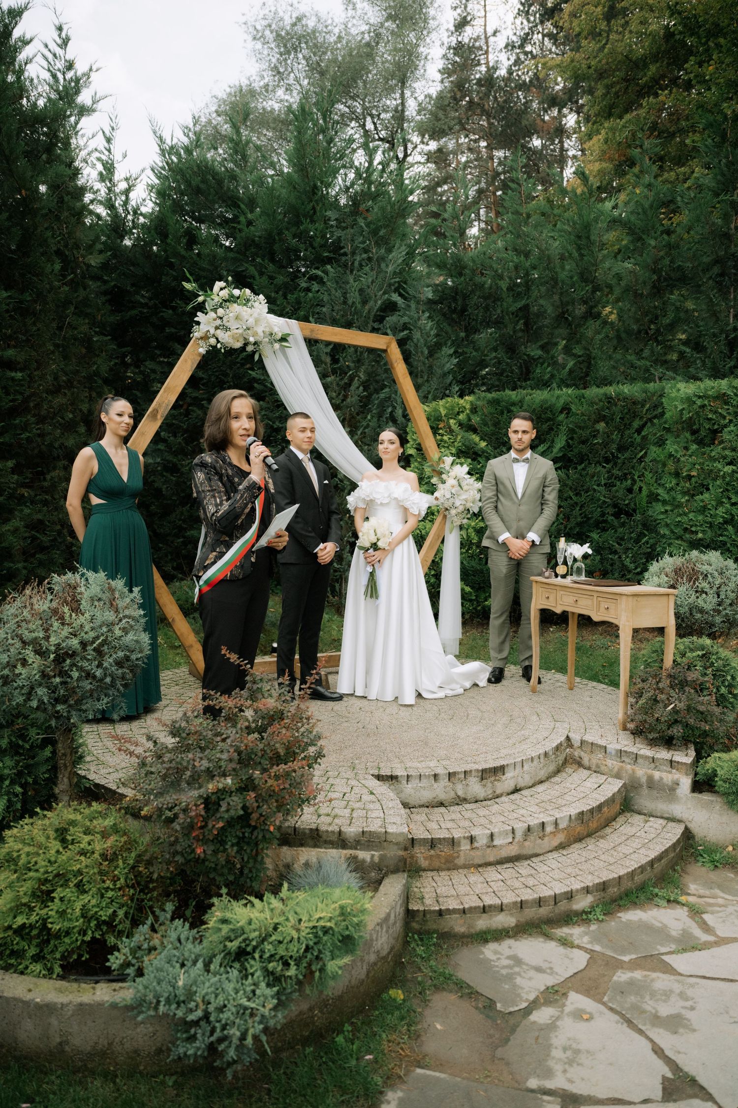 Wedding ceremony taking place at a garden venue with curved stone steps and lush greenery in the background.