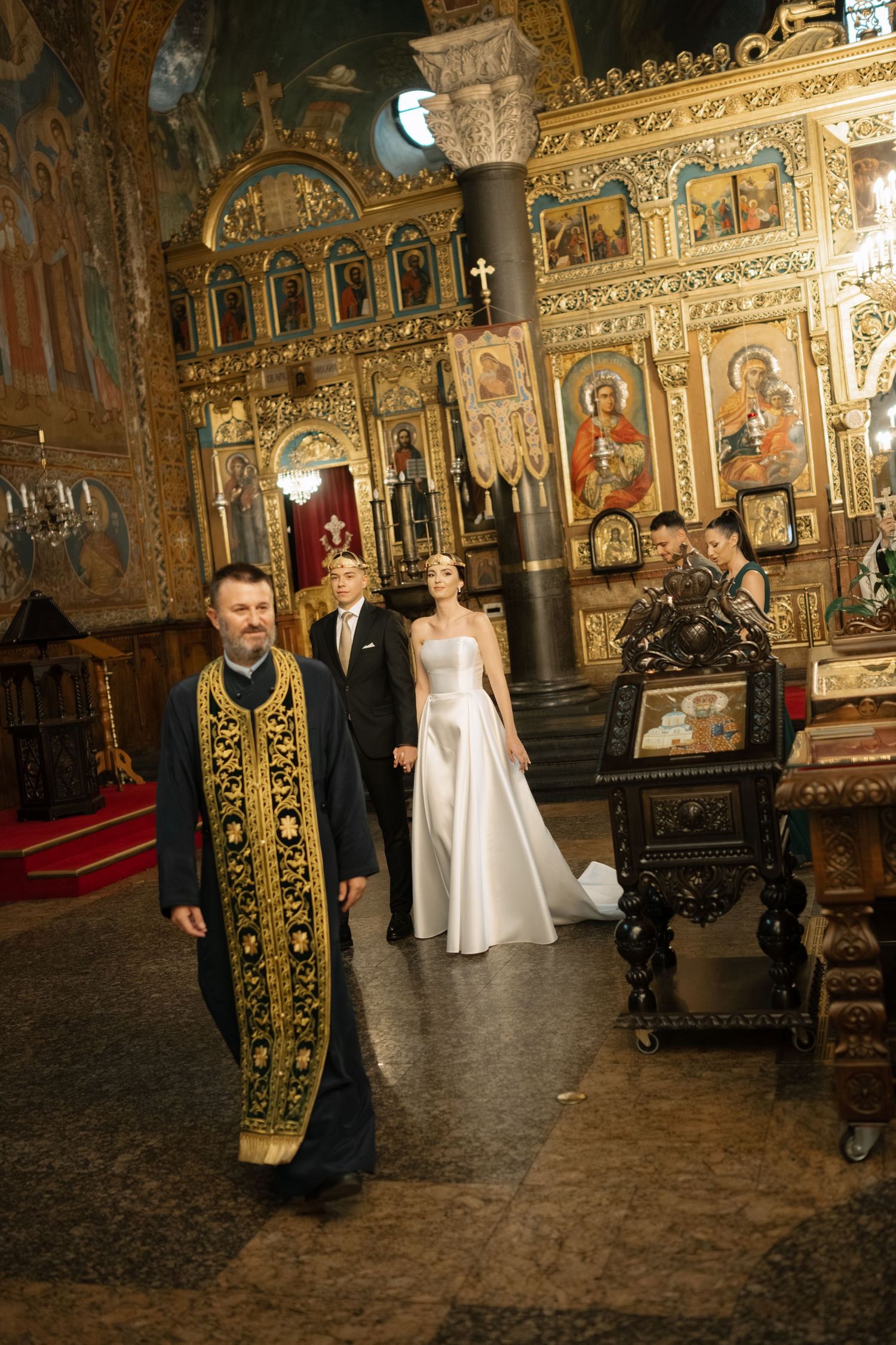 Orthodox wedding ceremony taking place inside an ornate church with gilded iconostasis and religious artwork decorating the walls.