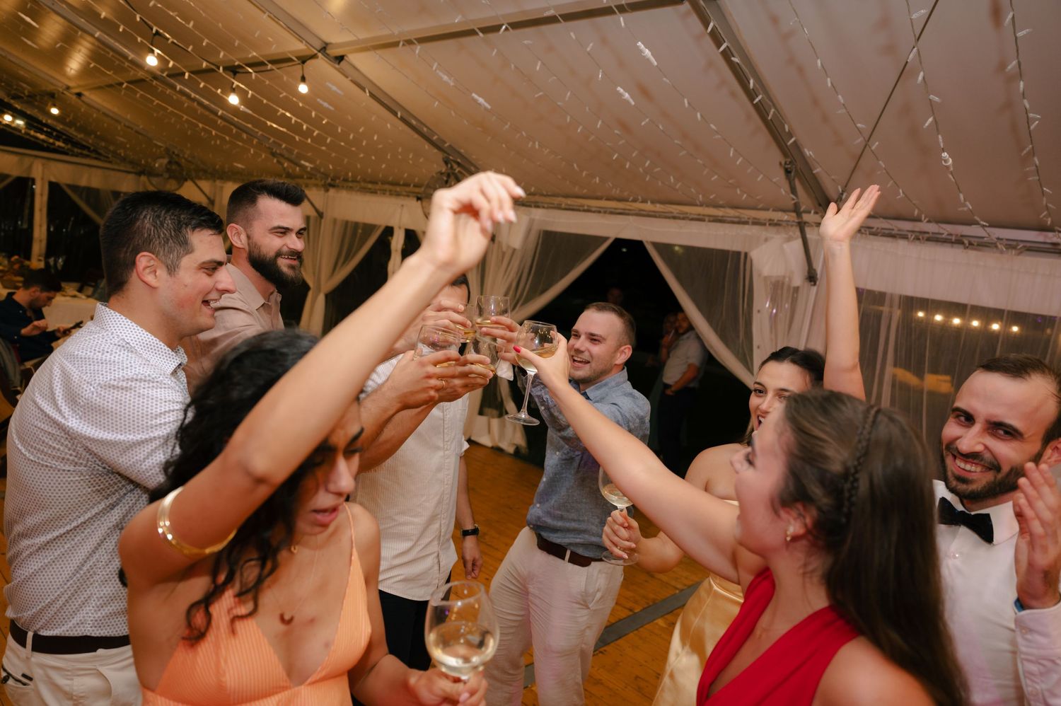 People dancing and celebrating at a lively indoor wedding reception with string lights overhead.