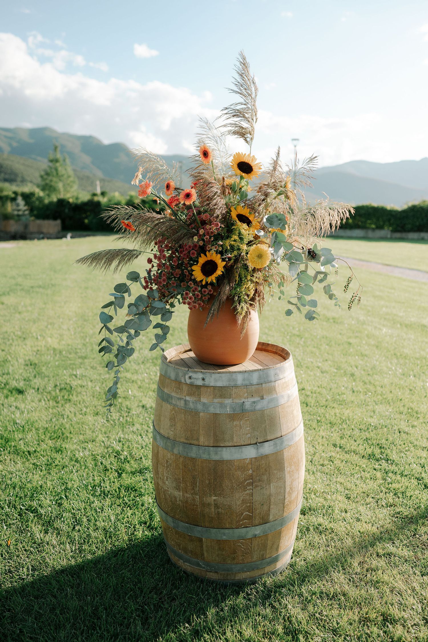 Rustic wooden wine barrel decorated with sunflowers and wildflowers on a grassy hillside at sunset.