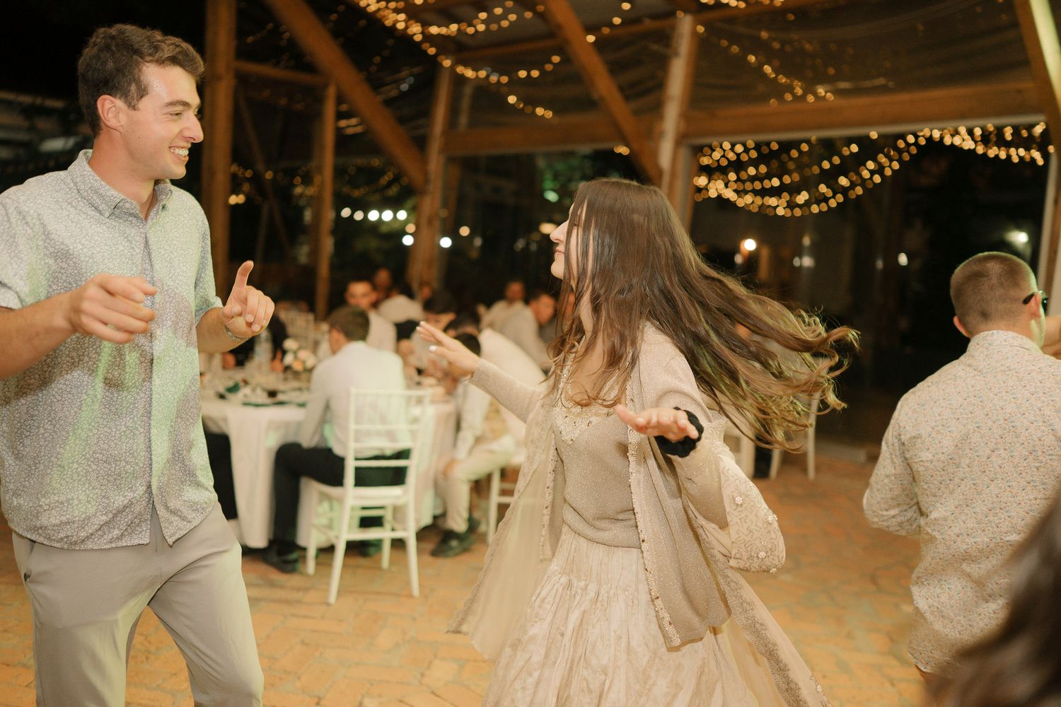 Wedding guests dance and celebrate on a rustic wooden dance floor illuminated by overhead string lights.