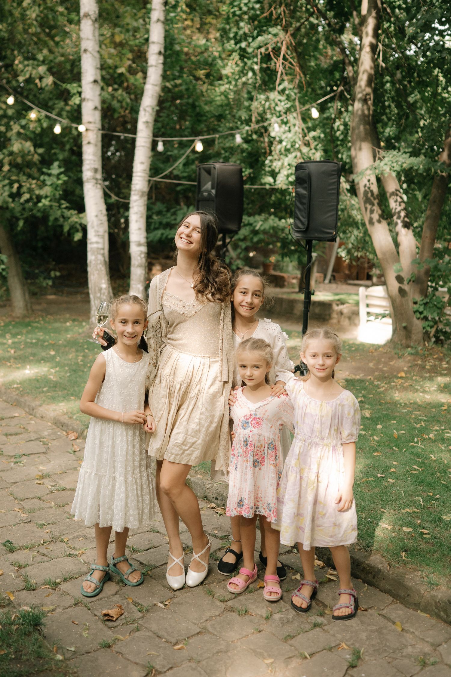 Young children in light colored dresses standing together on a stone pathway in a garden setting.