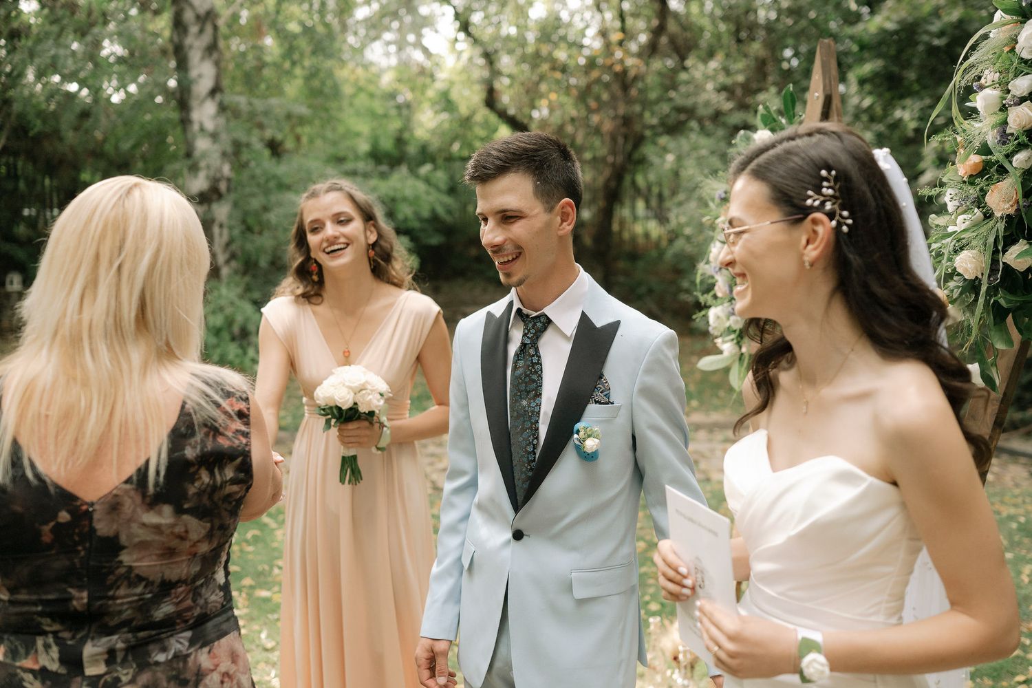 Wedding party in peach bridesmaid dresses and light blue suits celebrates in a lush garden setting.