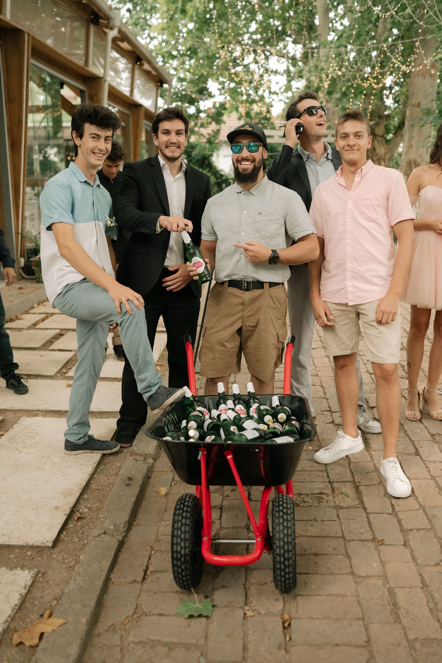 Group of friends gathered outdoors with red utility cart filled with beverages on brick pathway.