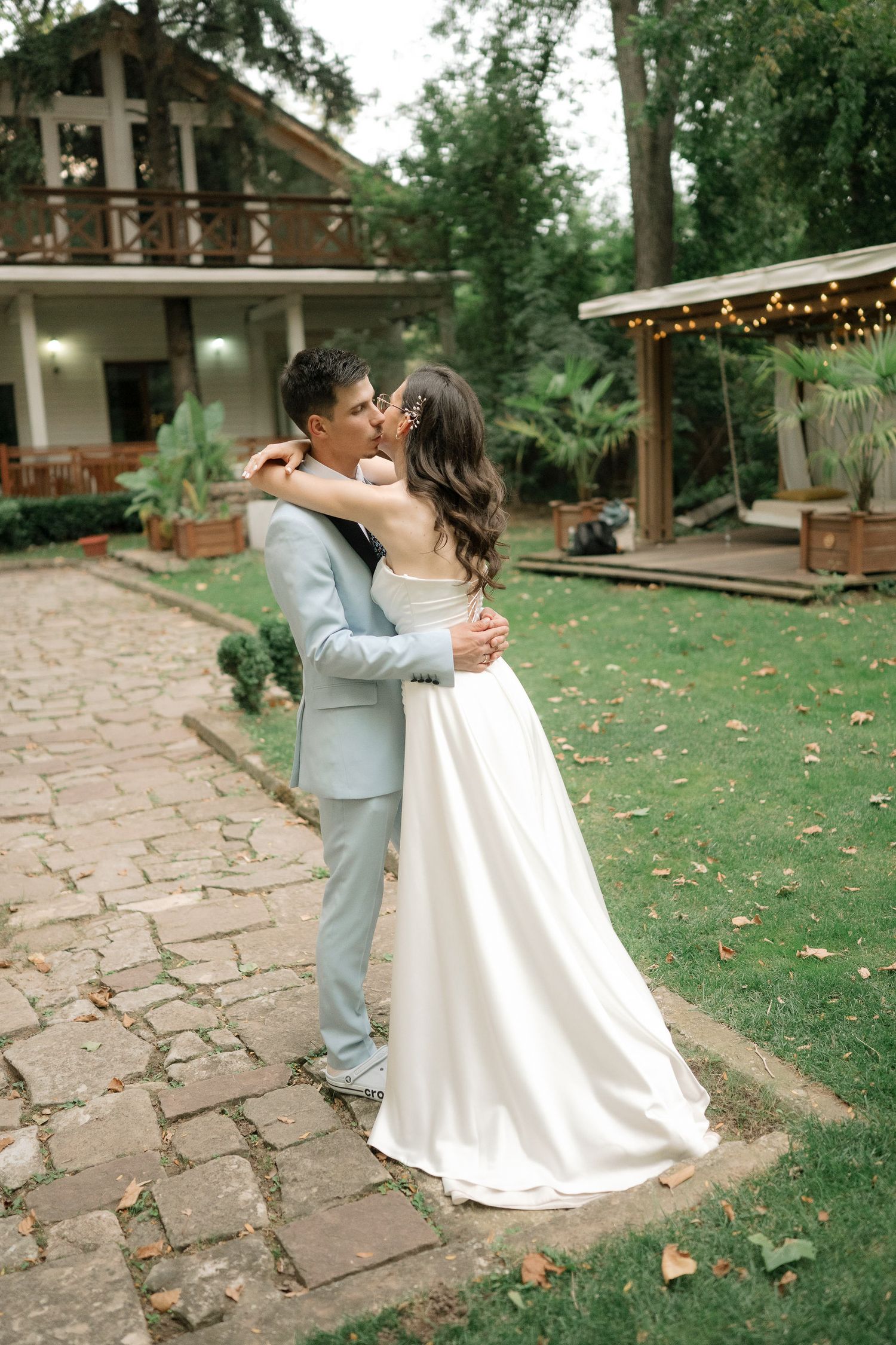 A couple in wedding attire shares a romantic dance on a stone patio surrounded by lush greenery and string lights.