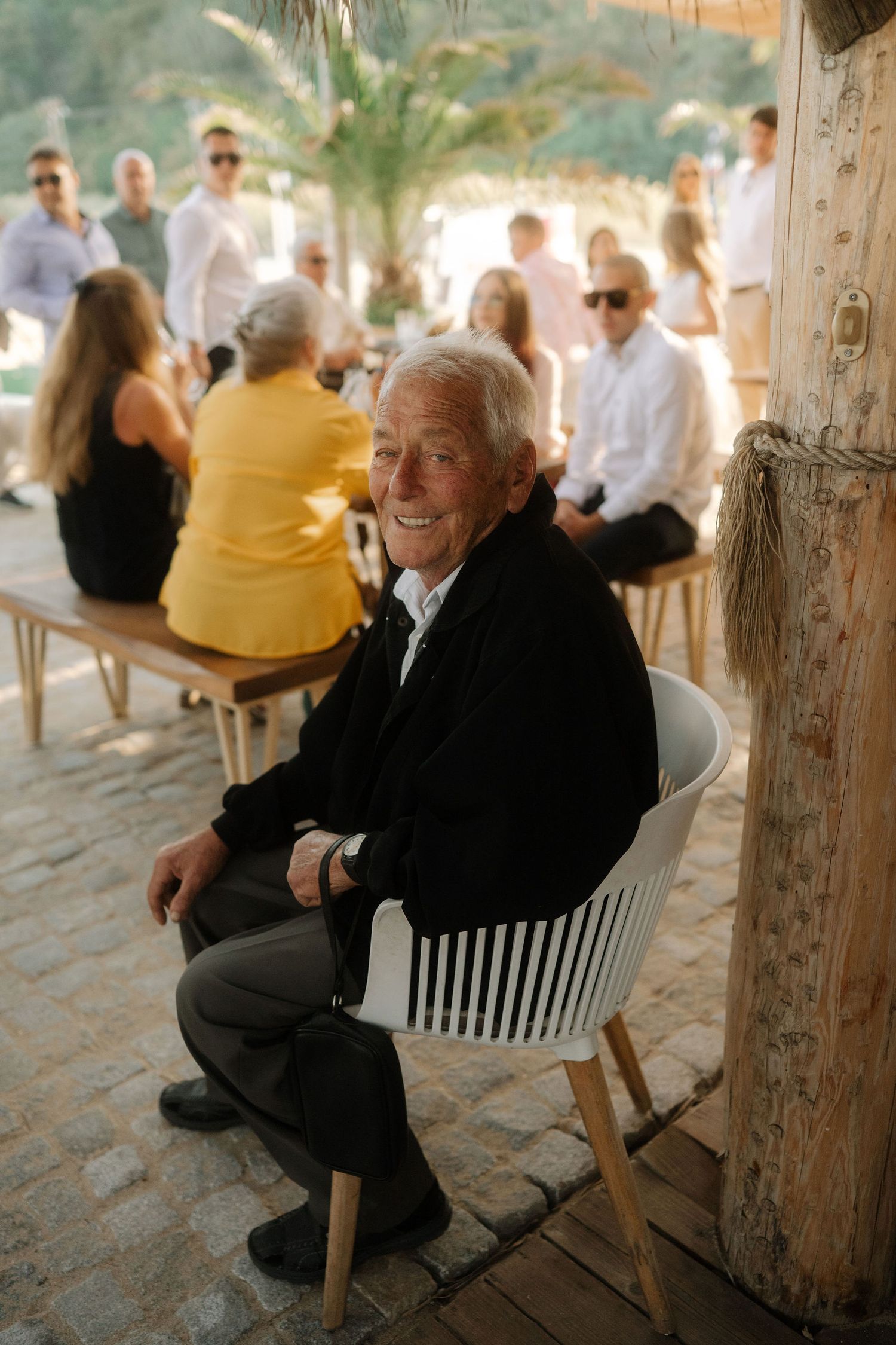 A person in a black suit sits and smiles warmly while relaxing on a covered porch area at an outdoor venue.