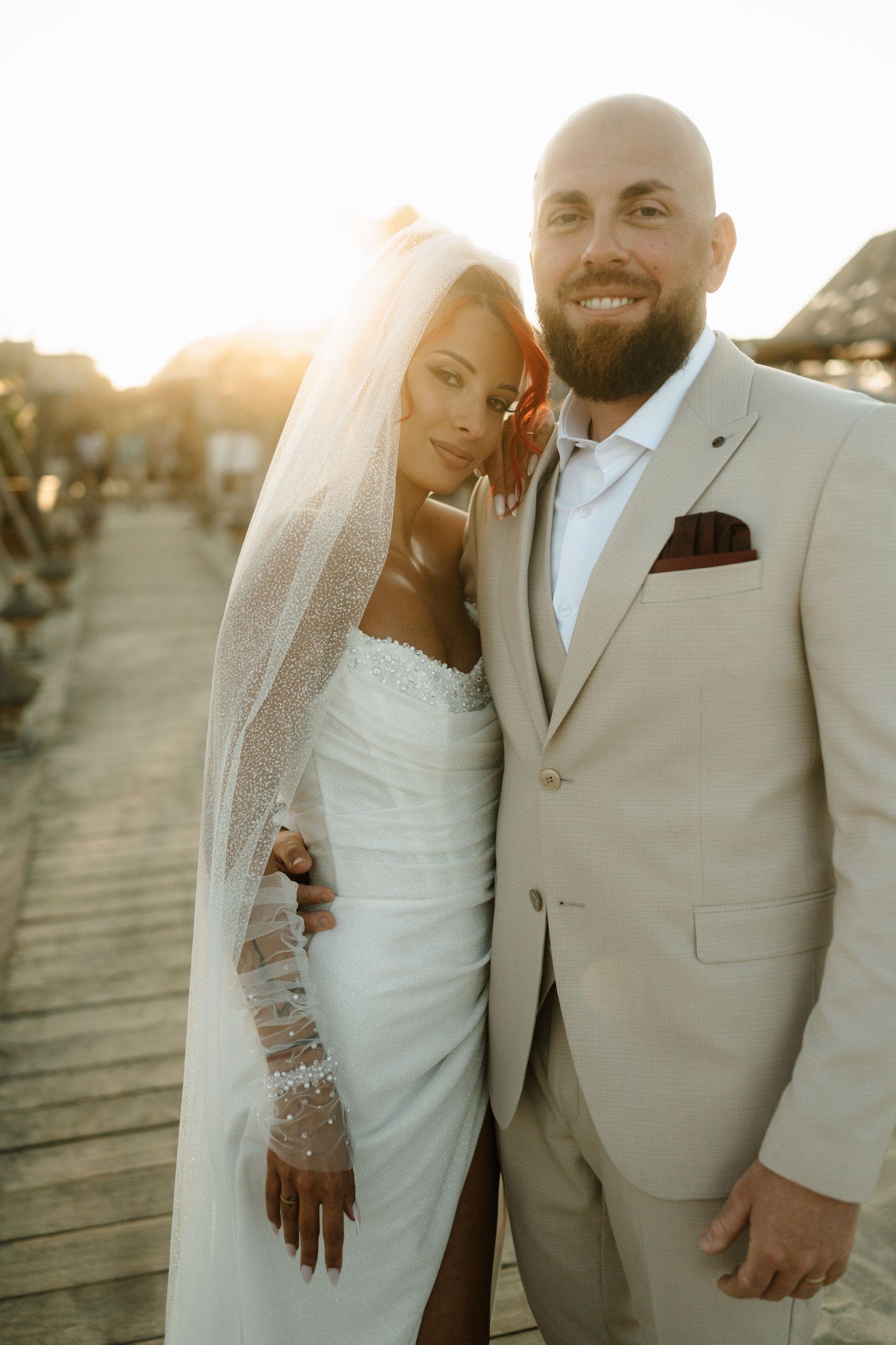 A happy couple in wedding attire poses together on a wooden boardwalk at sunset.
