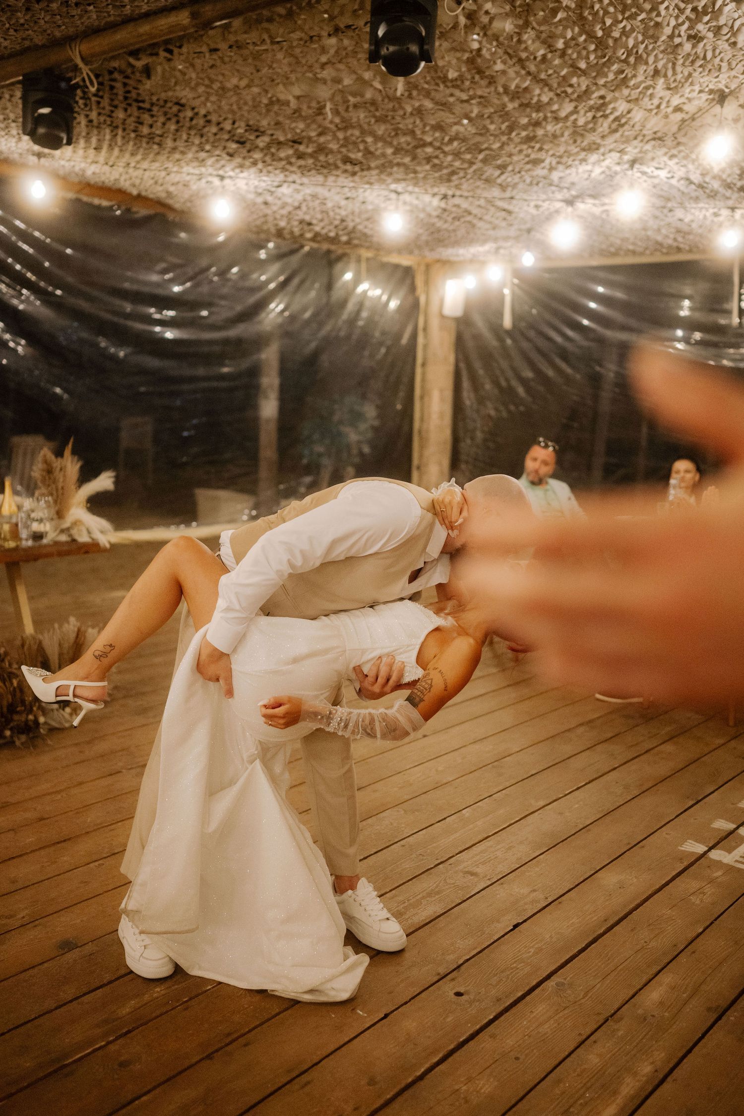A person in a white dress lies gracefully on a rustic wooden floor under string lights at an indoor venue.