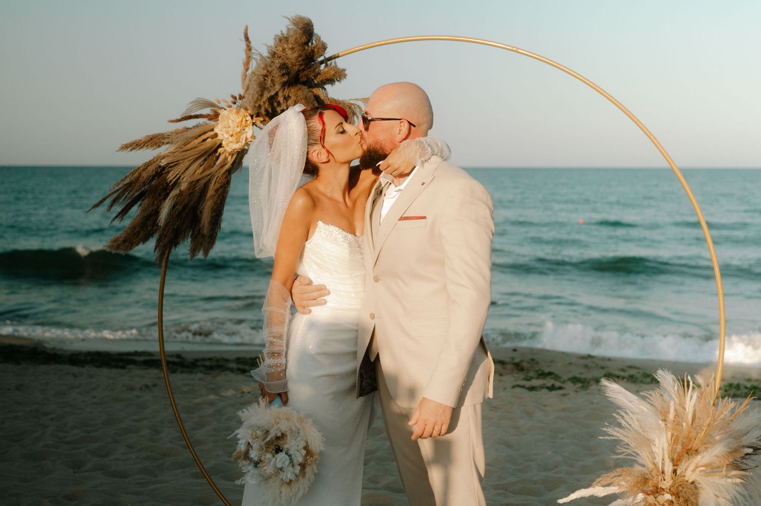 A couple embraces on a beach at sunset with dried pampas grass decorations and ocean waves in the background.