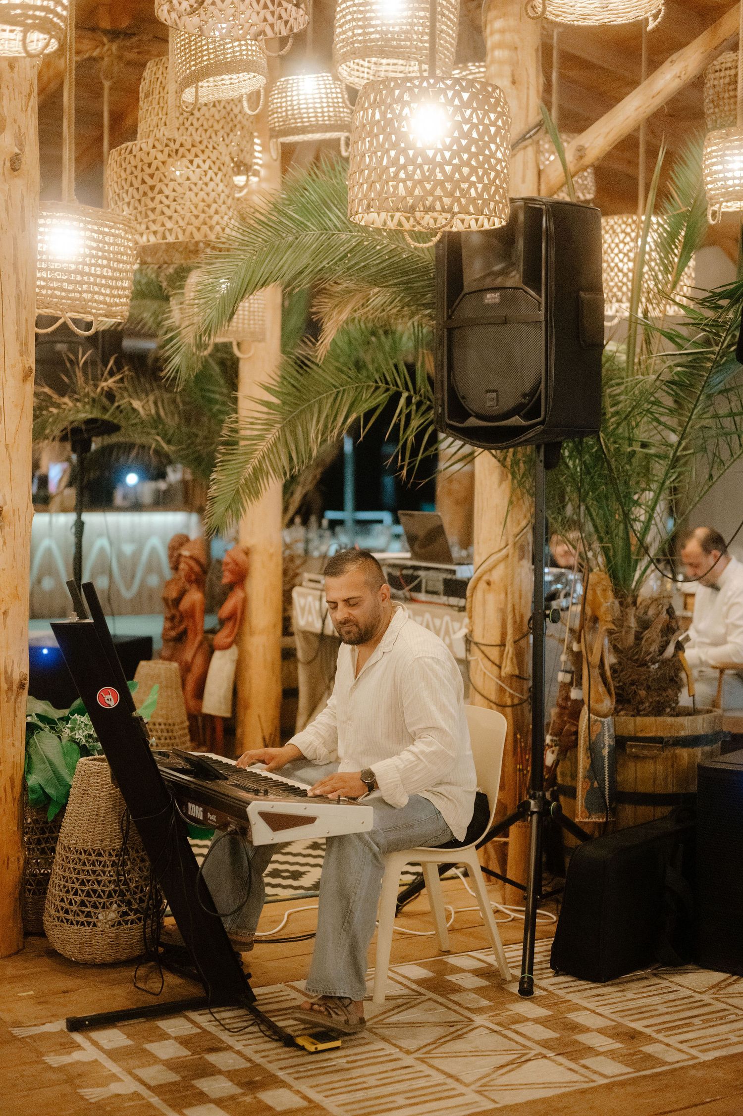 Musicians perform at a tropical-themed venue with woven lamp shades and palm plants creating an intimate ambiance.