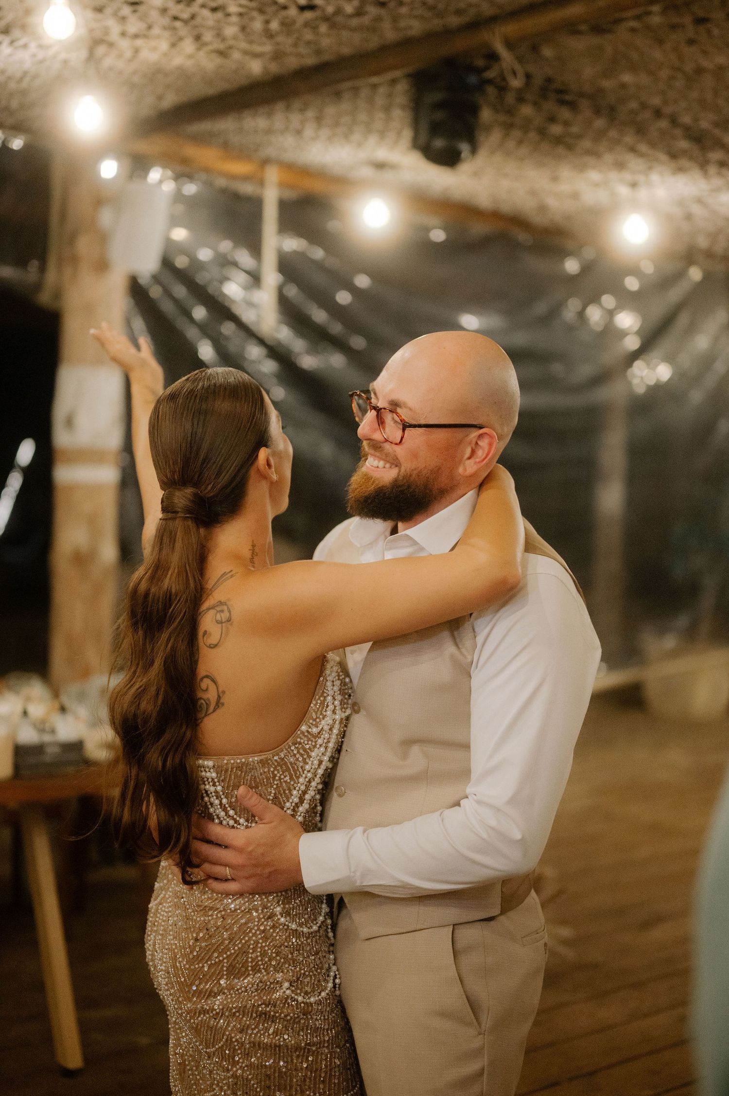 A couple shares an intimate dance together under warm lighting at a rustic indoor venue with string lights overhead.