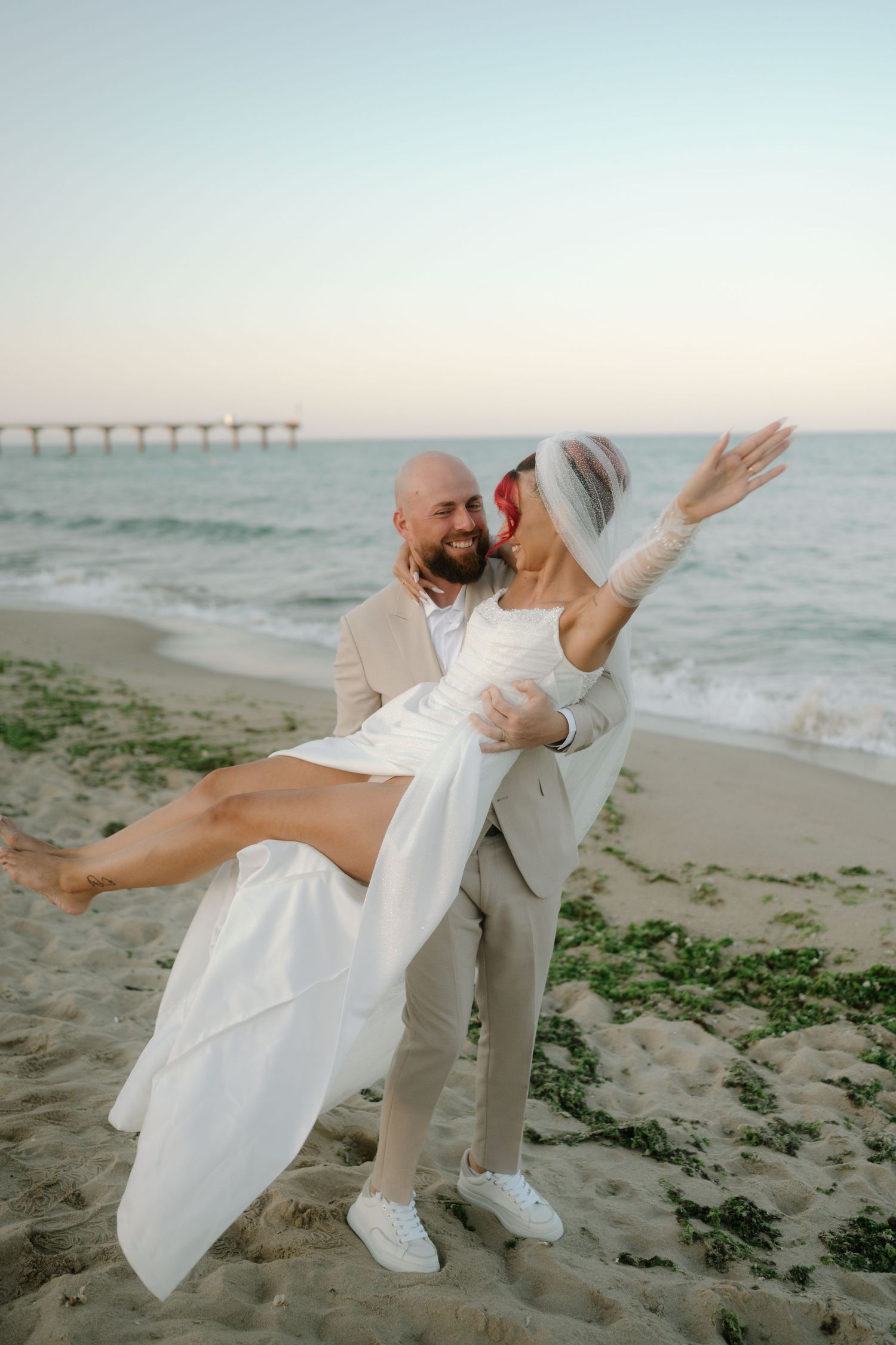 A couple shares a romantic moment dancing on the beach at sunset with a pier visible in the background.