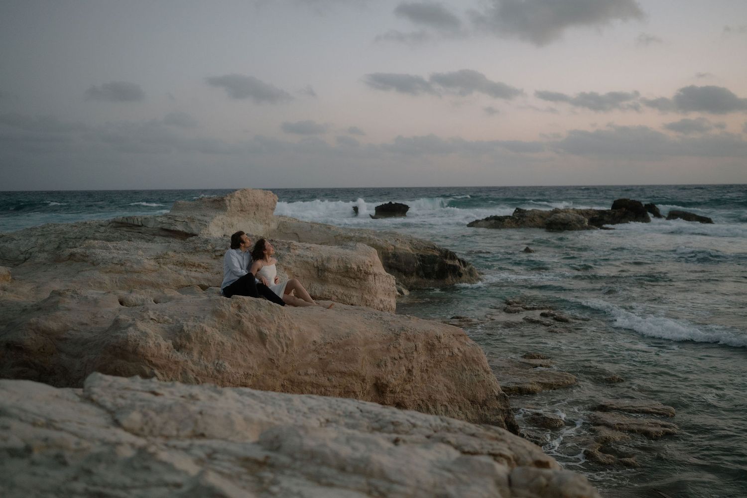 Couple sitting on rocky shoreline watching waves crash at sunset with dramatic cloudy sky.