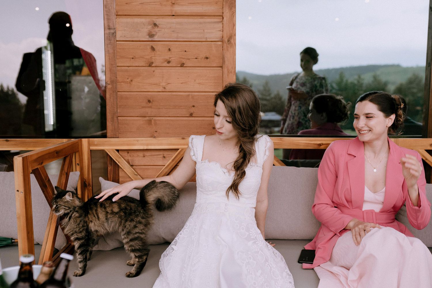 A tabby cat sits on a bench next to wedding guests at a rustic outdoor venue with mountain views in the background.