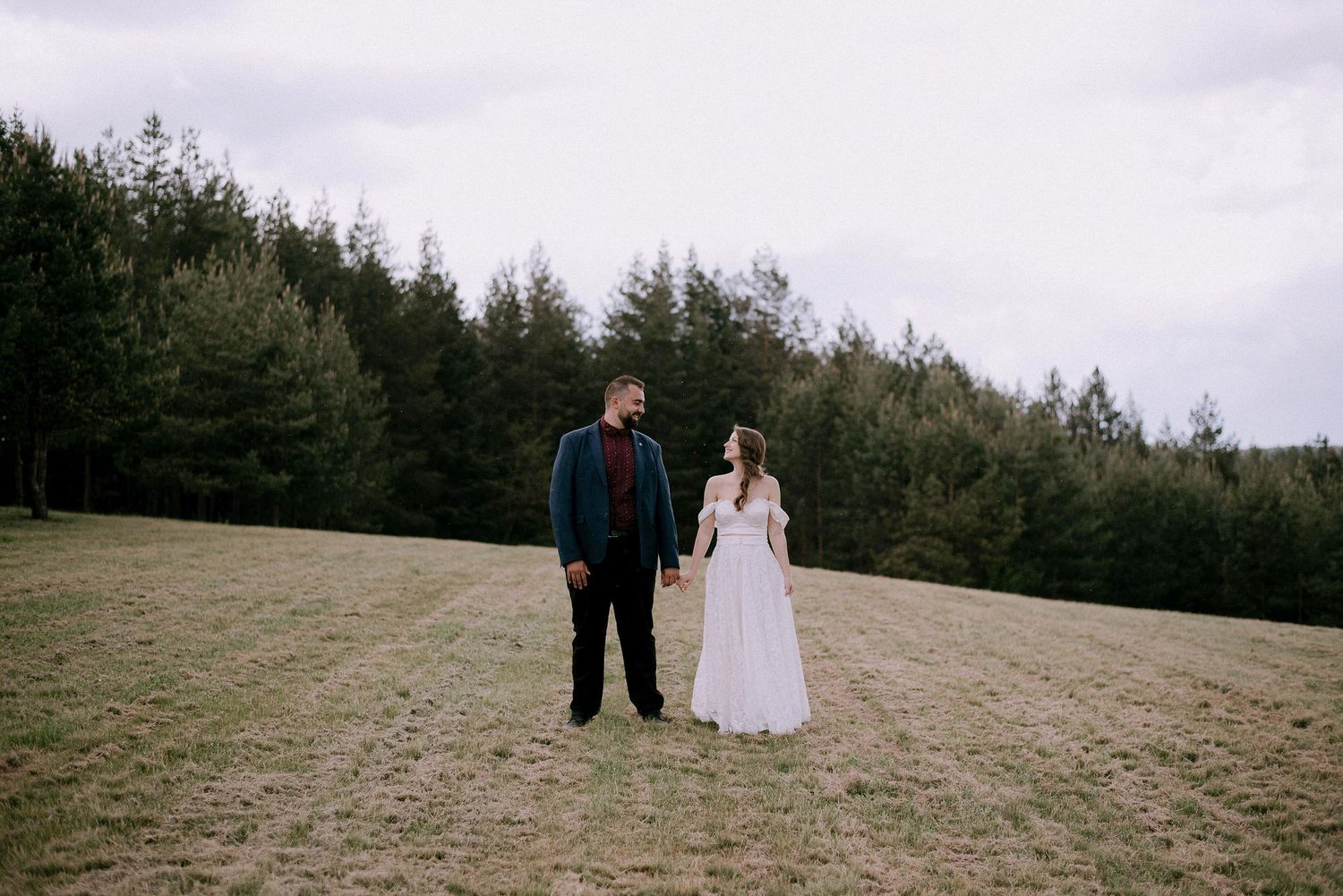 A couple in wedding attire stands together on a grassy hillside.