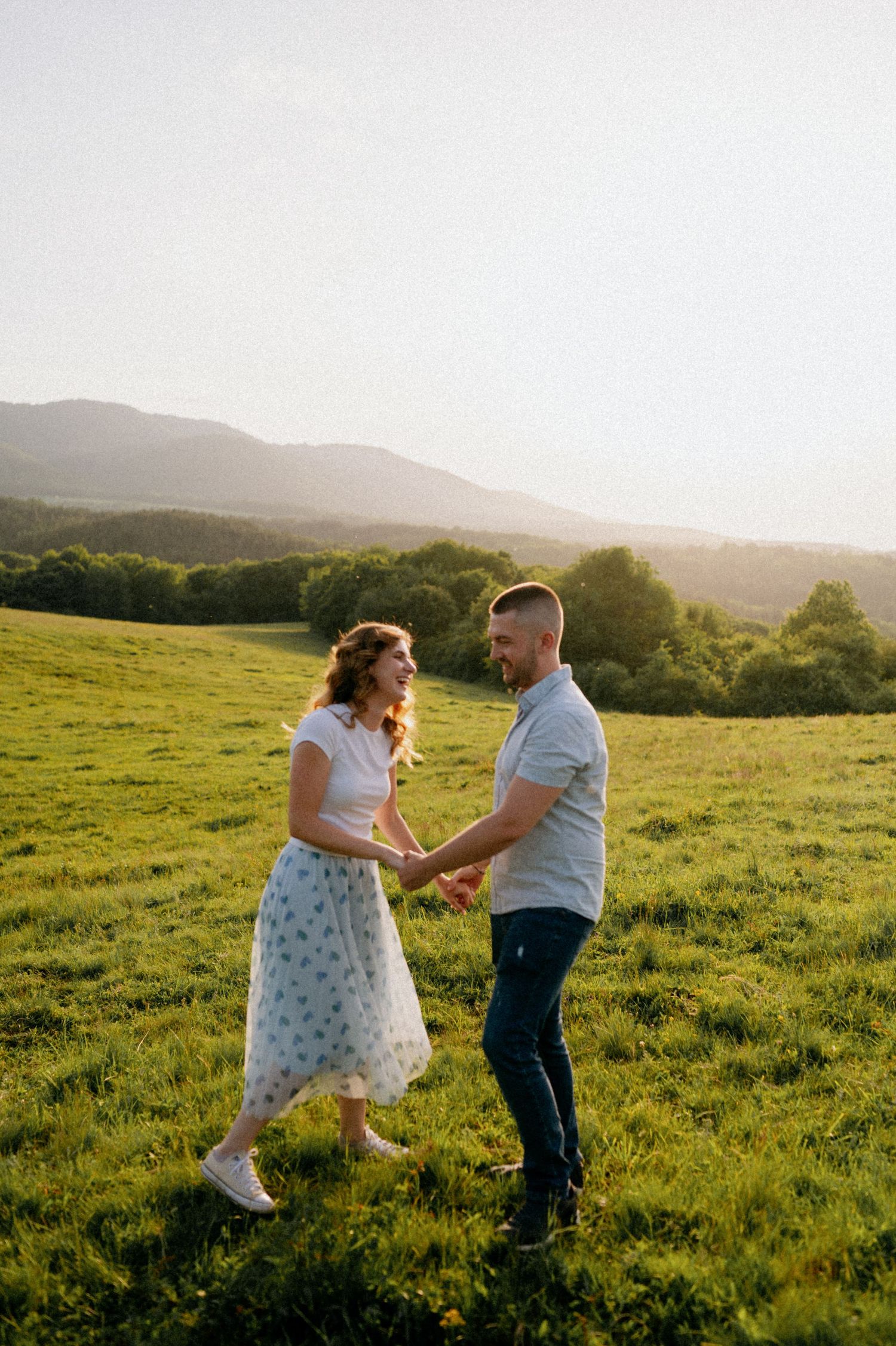 A couple shares a playful dance in a grassy field at sunset with rolling hills behind them.