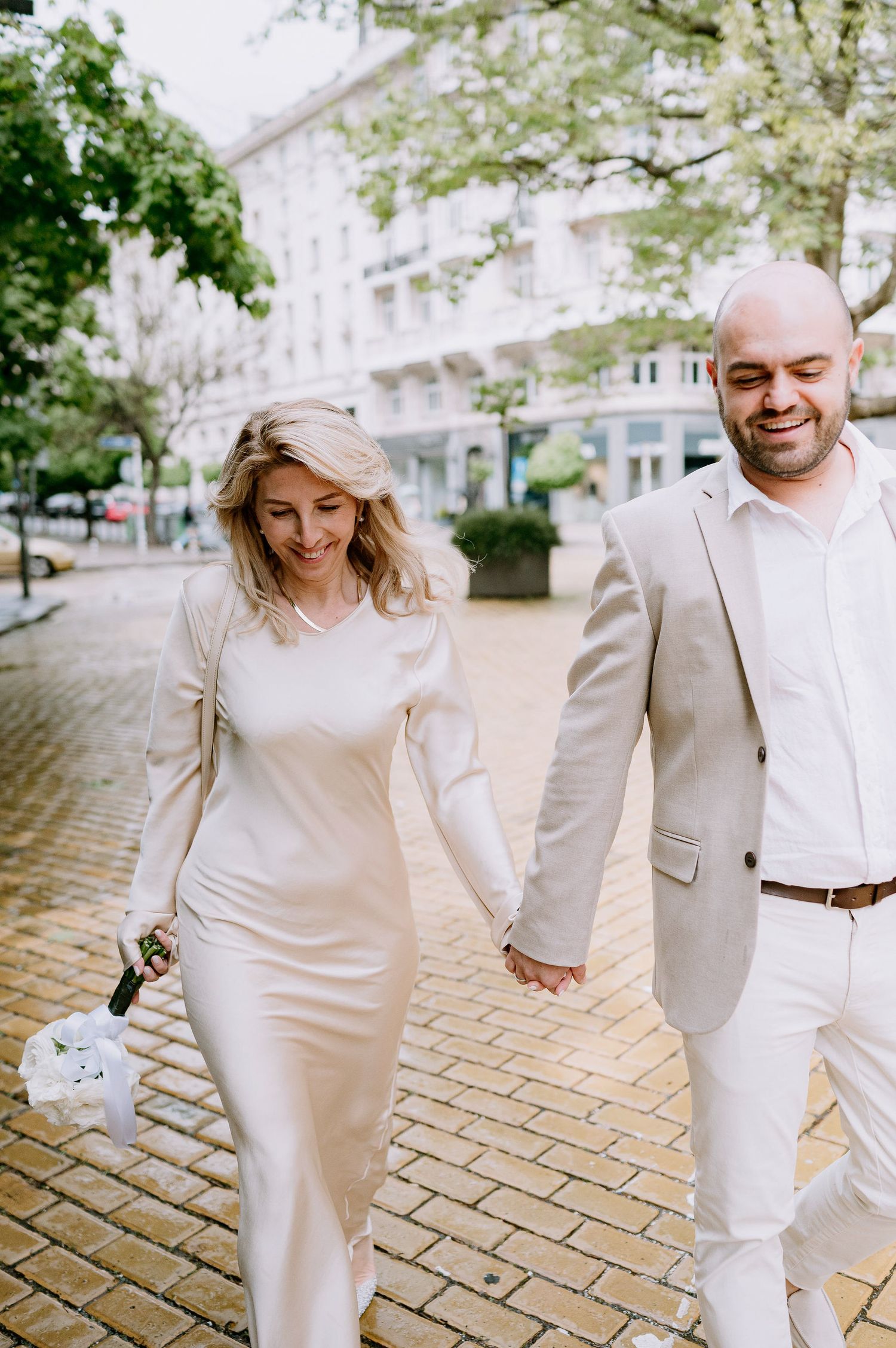 Couple in matching light-colored outfits walking and laughing together on cobblestone pavement.