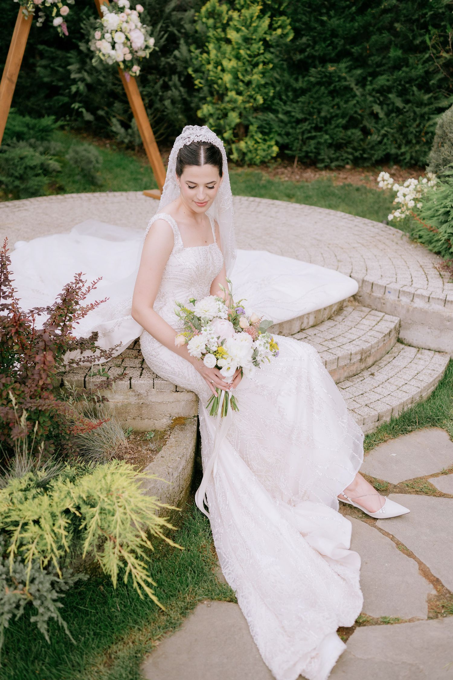 Bride in white dress poses on stone garden steps surrounded by ornamental grasses and flowers.