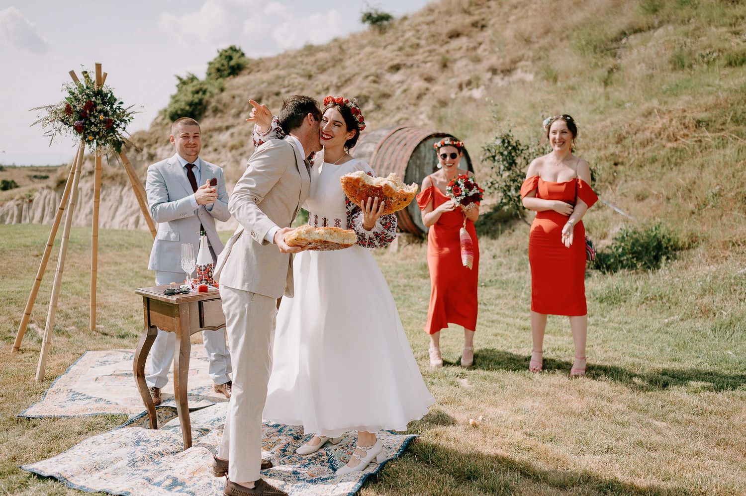 Wedding celebration on a sunny hillside with red-dressed bridesmaids and rustic wooden decor.