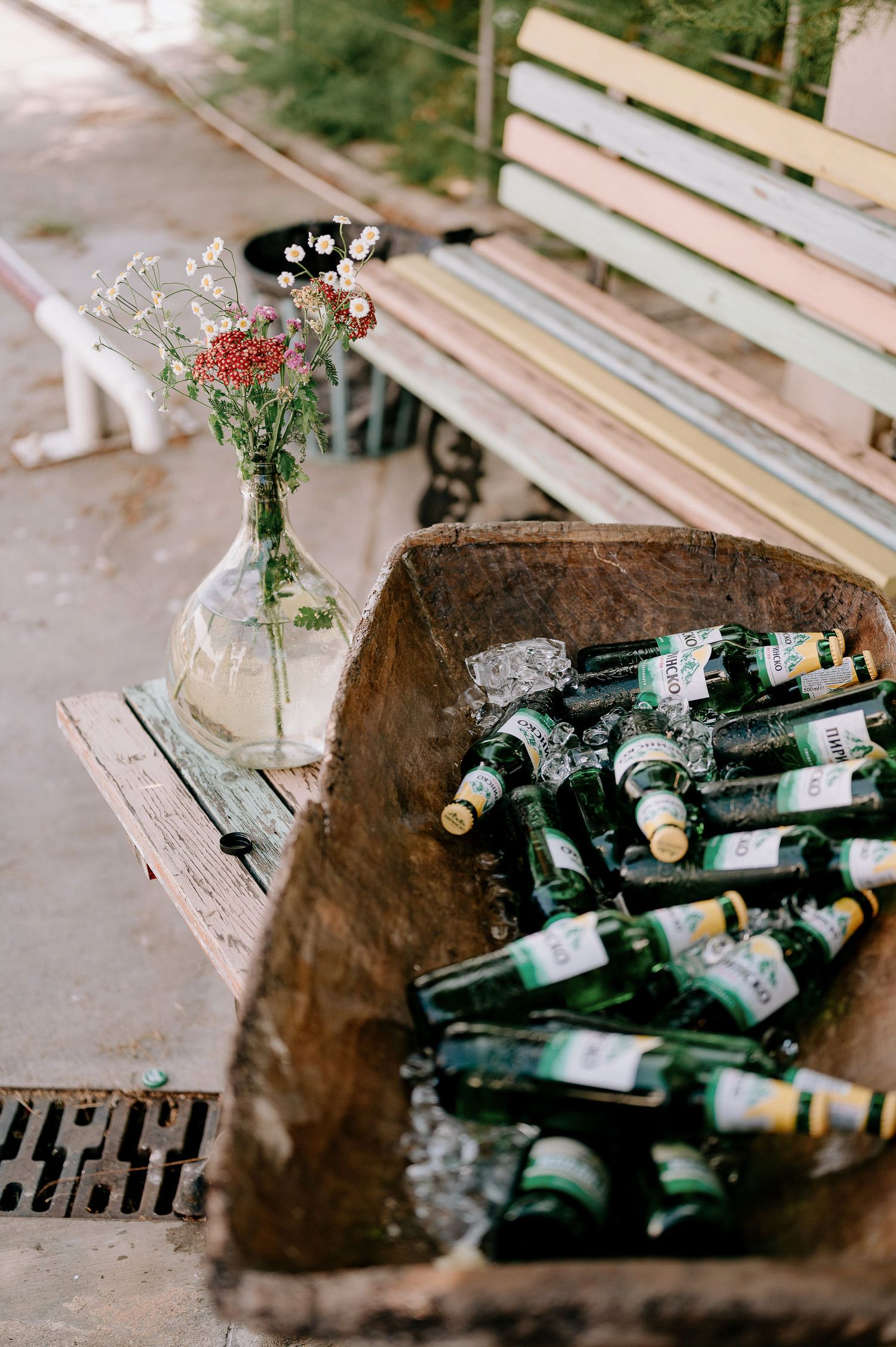 A rustic wooden trough filled with green wine bottles next to a delicate flower vase on a bench.