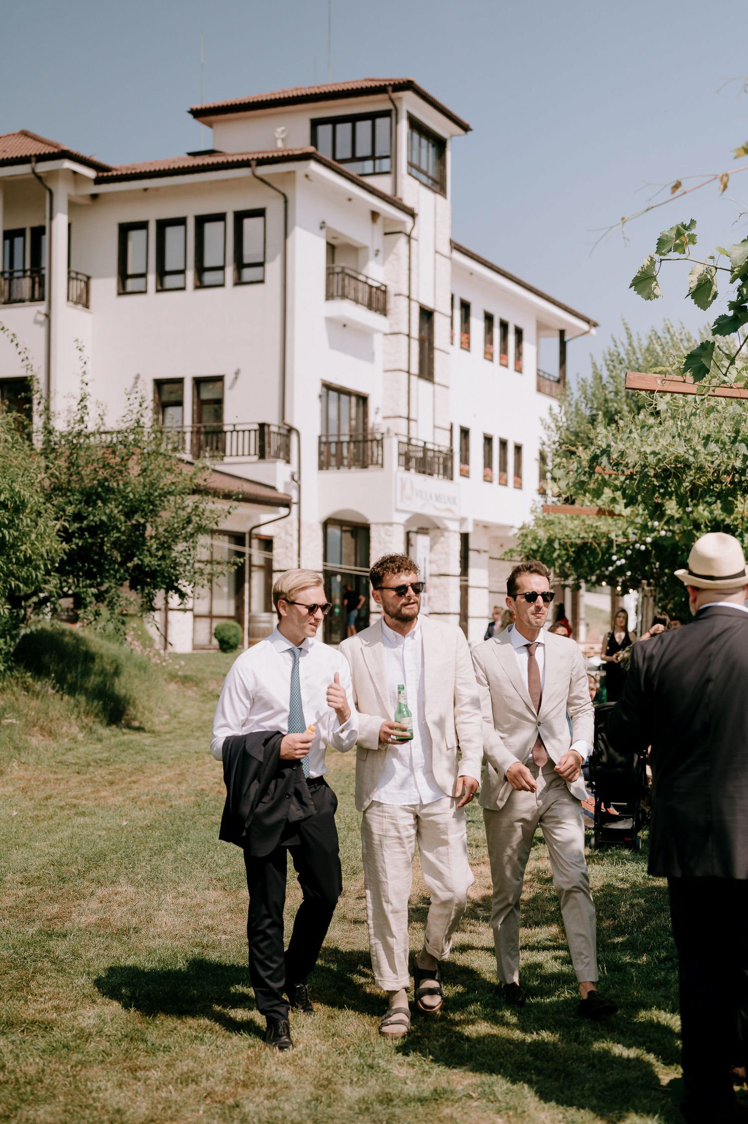 Groomsmen in light colored suits gather outside a white Mediterranean-style building.
