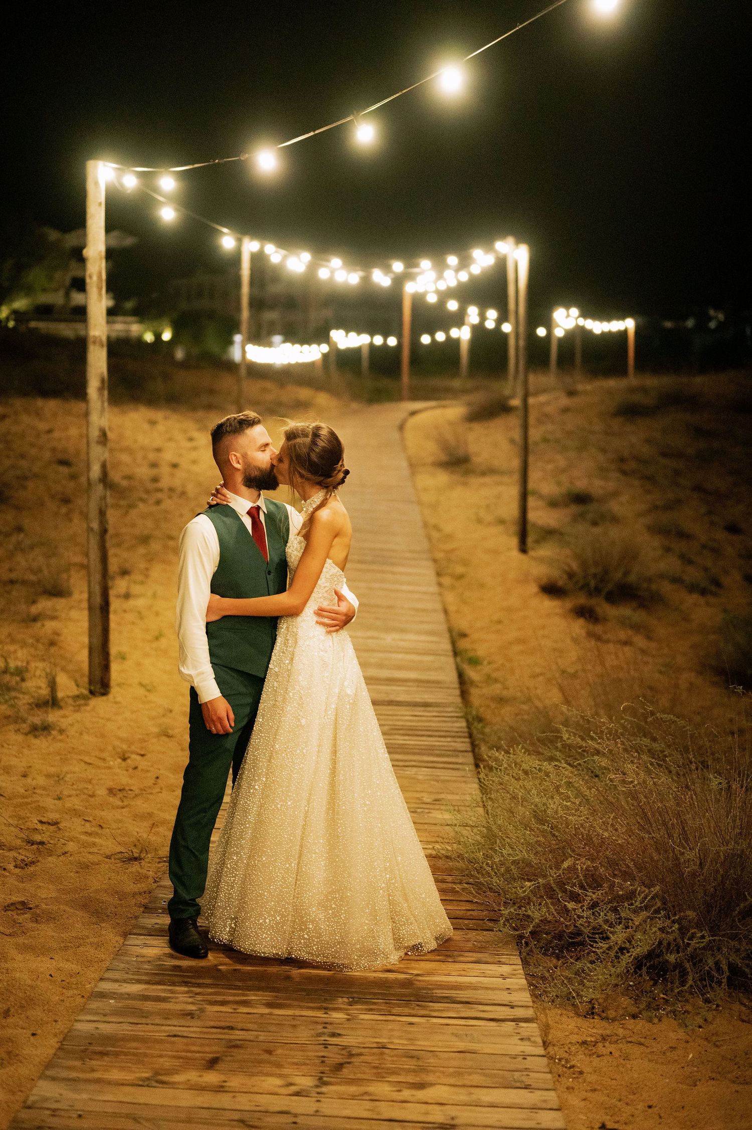 Romantic nighttime wedding photos on illuminated wooden boardwalk in desert setting.