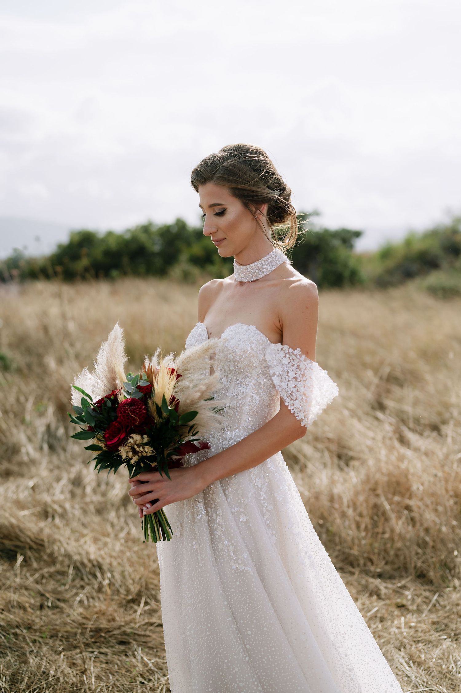 Side view of a bride in a romantic off-shoulder gown holding a deep red and green wedding bouquet.